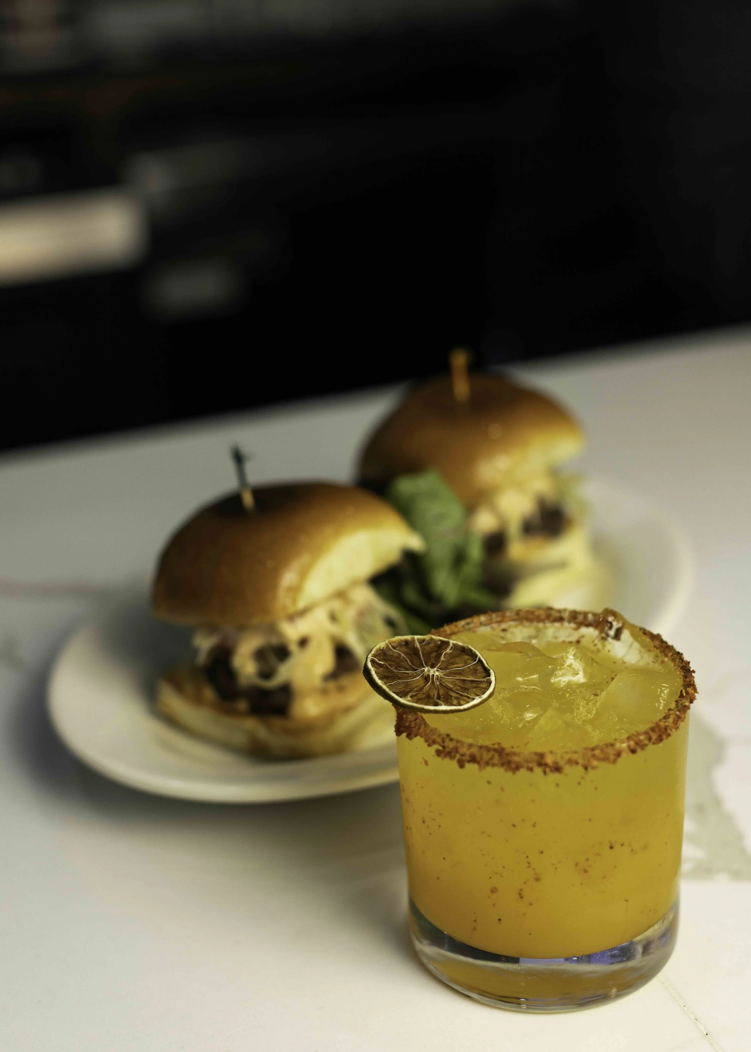 A yellow cocktail with a salted rim and a dried lime slice, served with two sliders with buns, cheese, lettuce, and meat, on a white plate.