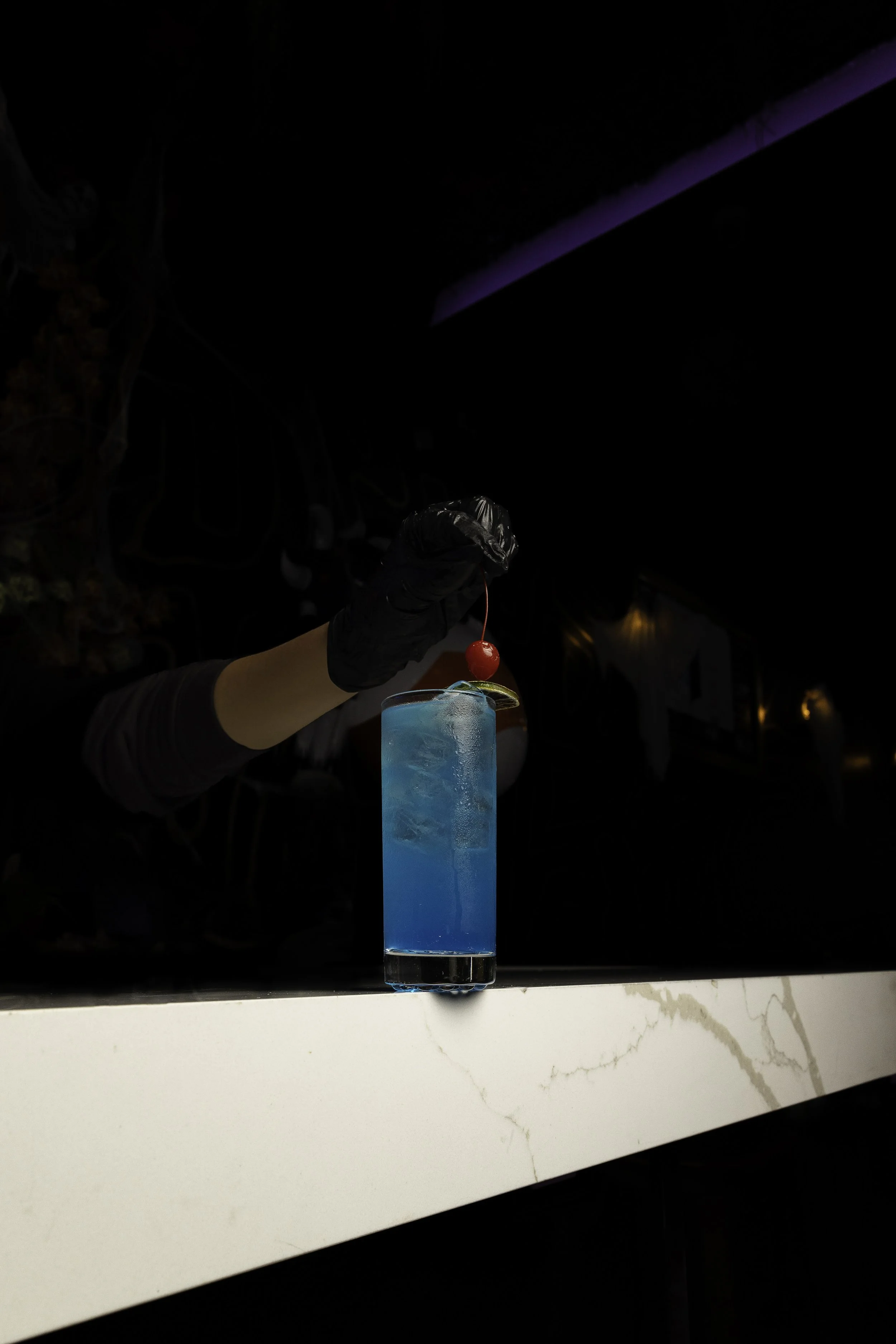 A bartender's hand in a black glove is garnishing a tall blue cocktail with a maraschino cherry and lime wedge in a dark bar setting with purple lighting.