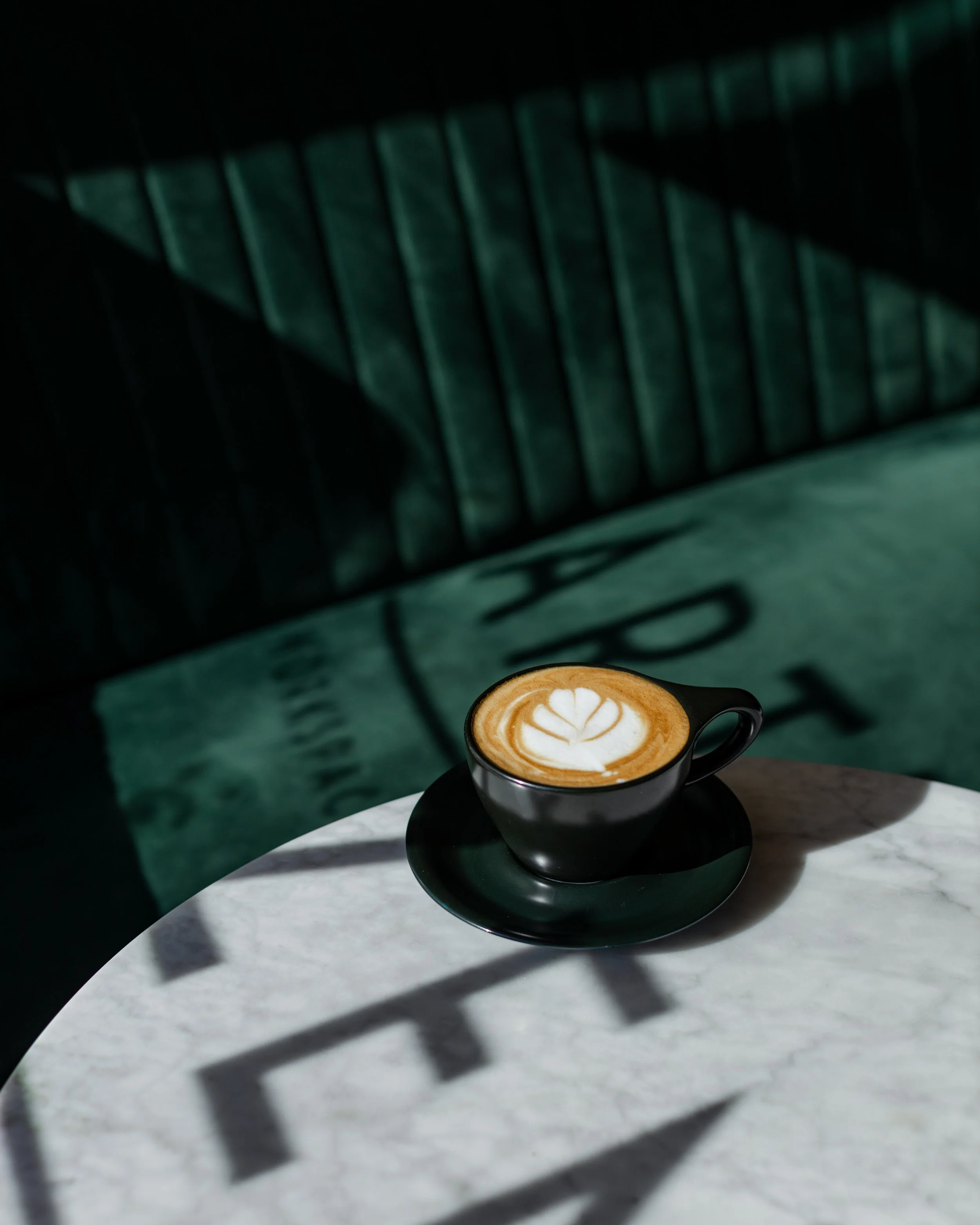 A black cup of coffee with latte art on a white marble table, with shadows and natural light creating patterns.