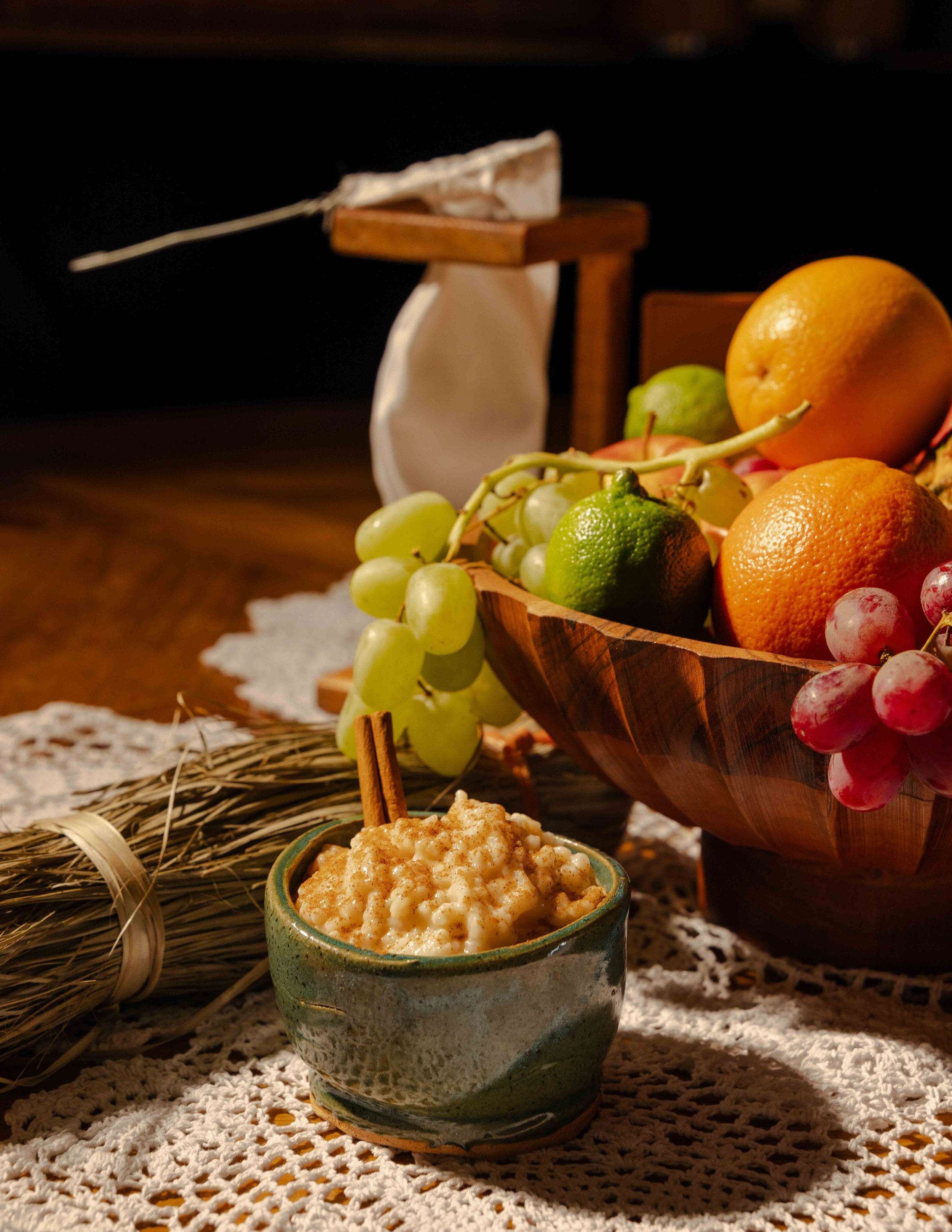 A bowl of rice pudding with cinnamon and a cinnamon stick in the foreground. A bunch of green grapes, a lime, oranges, a pear, and red grapes are in a wooden bowl behind it. A bundle of wheat and a small sculpture of a bag or pouch are also on the la