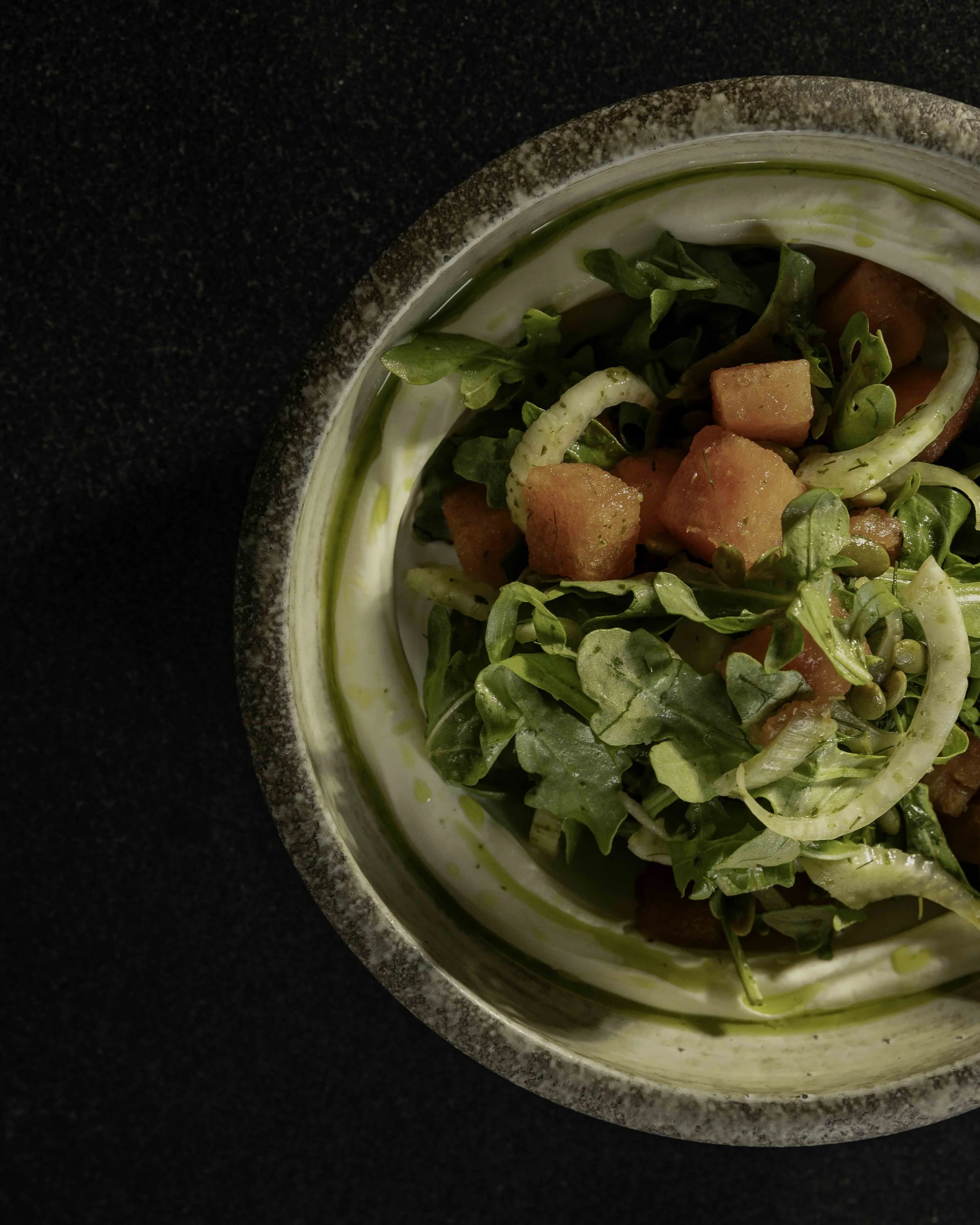 Close-up of a salad in a ceramic bowl, containing watermelon pieces, leafy greens, and sliced onions on a black surface.