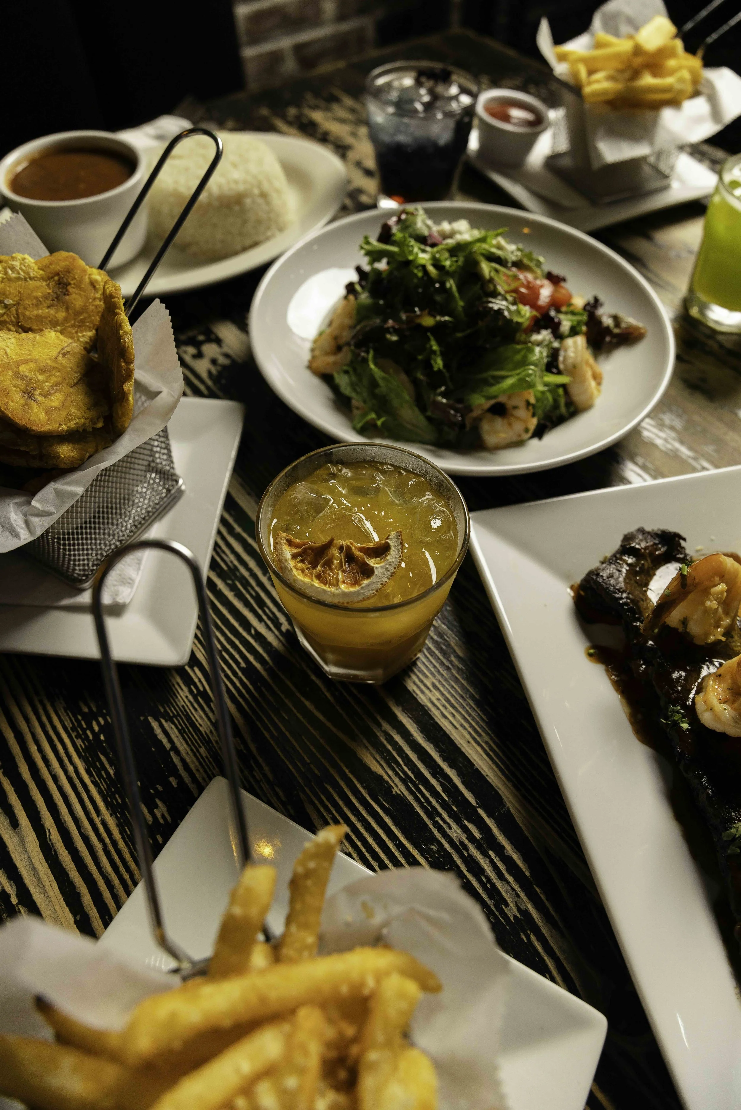 Assorted dishes on a rustic wooden table including fries, a salad, a glass of iced tea with lemon, a barbecue dish, seasoned grilled chicken, rice, and dipping sauces.