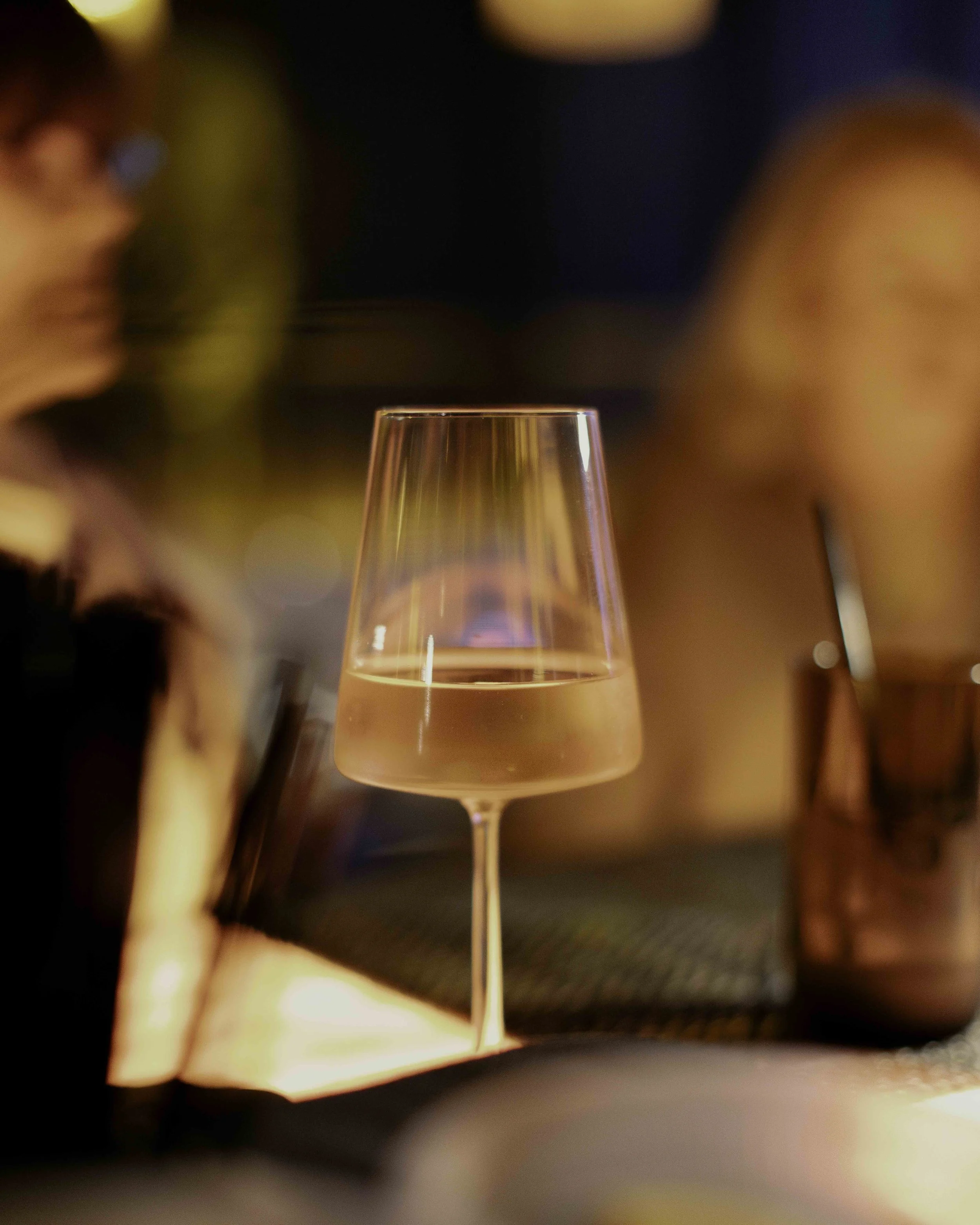 A glass of rosé wine on a table in a dimly lit setting, with blurred people in the background.