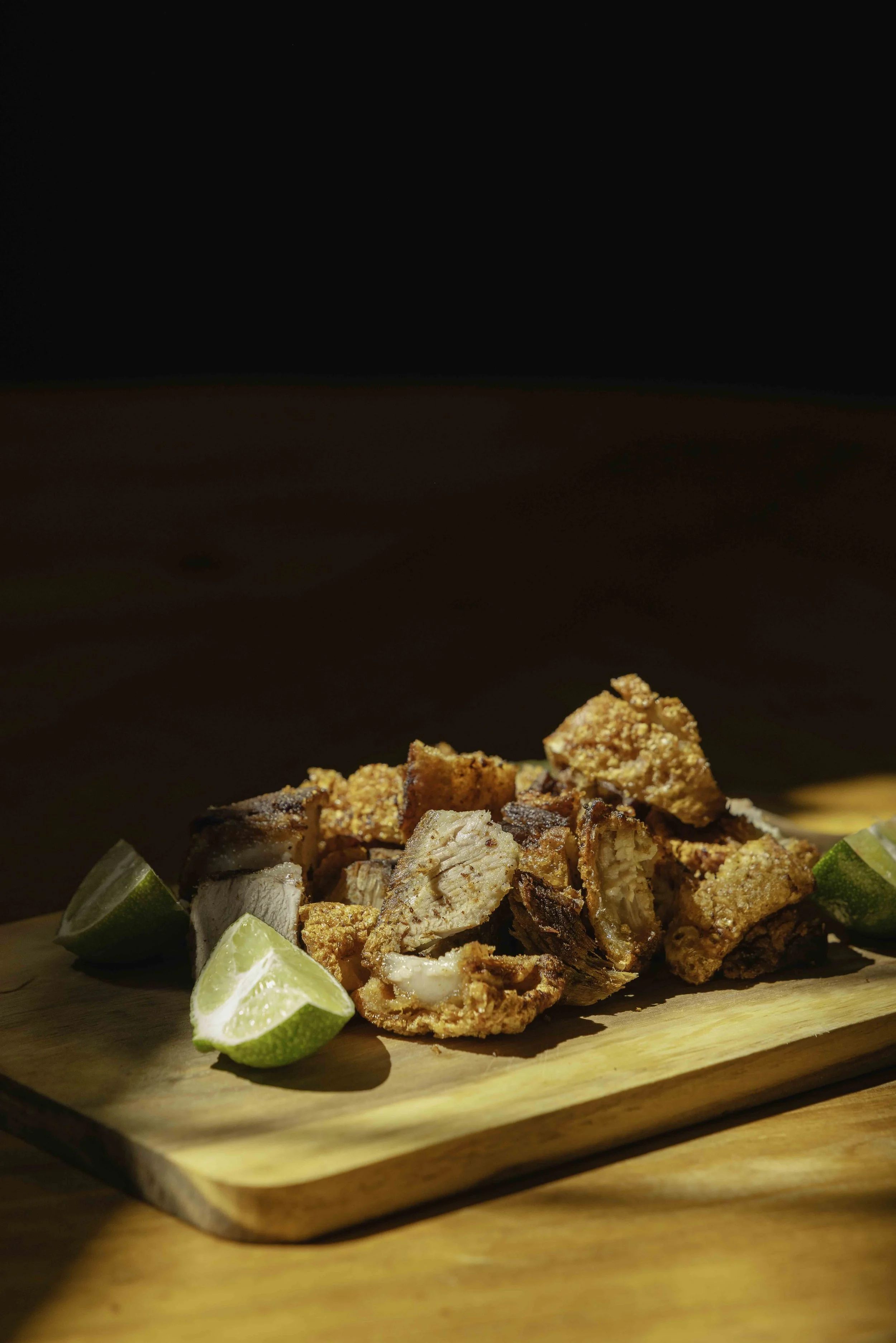 Fried pork belly slices with lime wedges on a wooden cutting board.