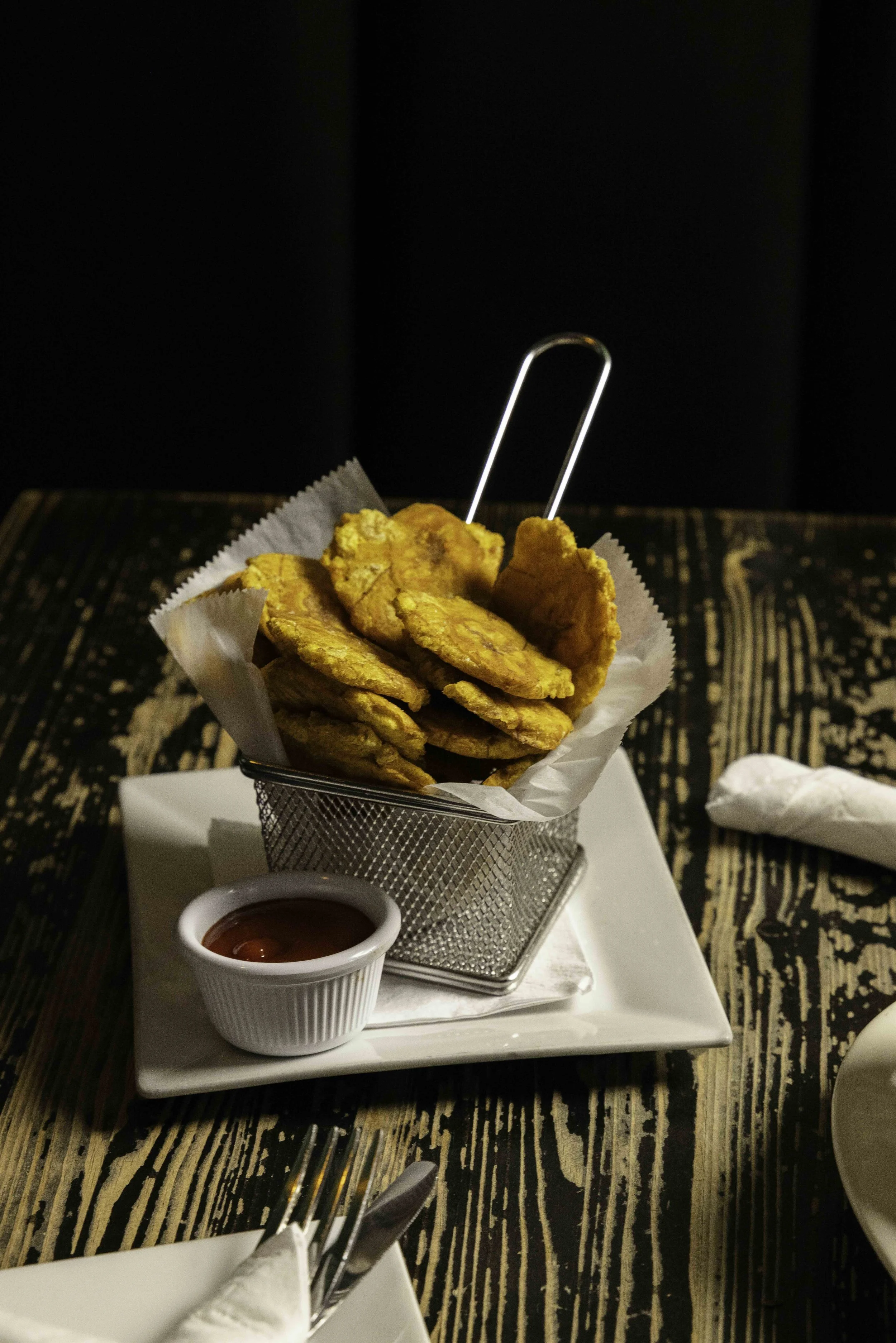 Basket of fried plantains with a side of ketchup, served on a white plate on a rustic wooden table.