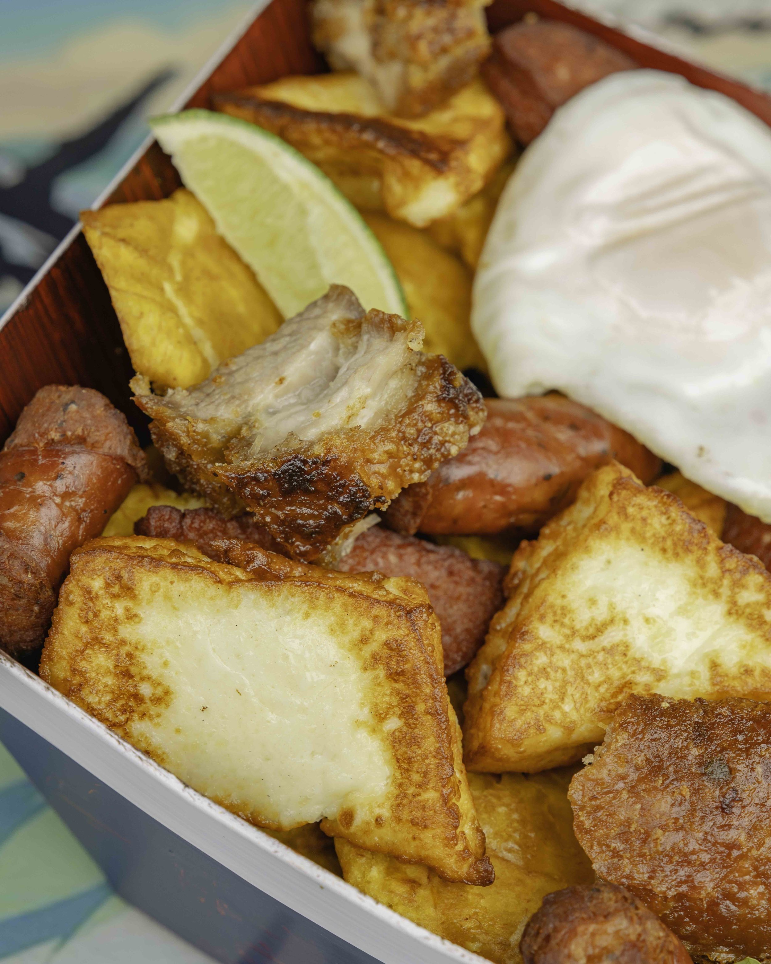 A close-up of a savory breakfast platter with fried potatoes, sausage, bacon, a poached egg, grilled vegetable, and a lemon wedge.