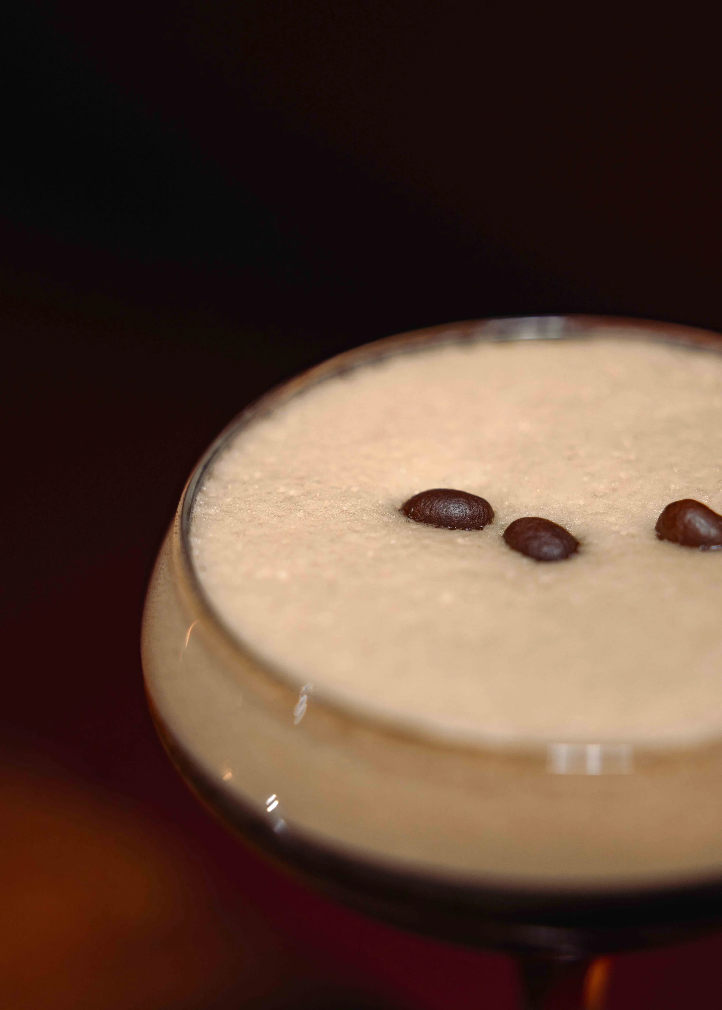 Close-up of a creamy coffee drink with three coffee beans on top in a glass mug against a dark background.