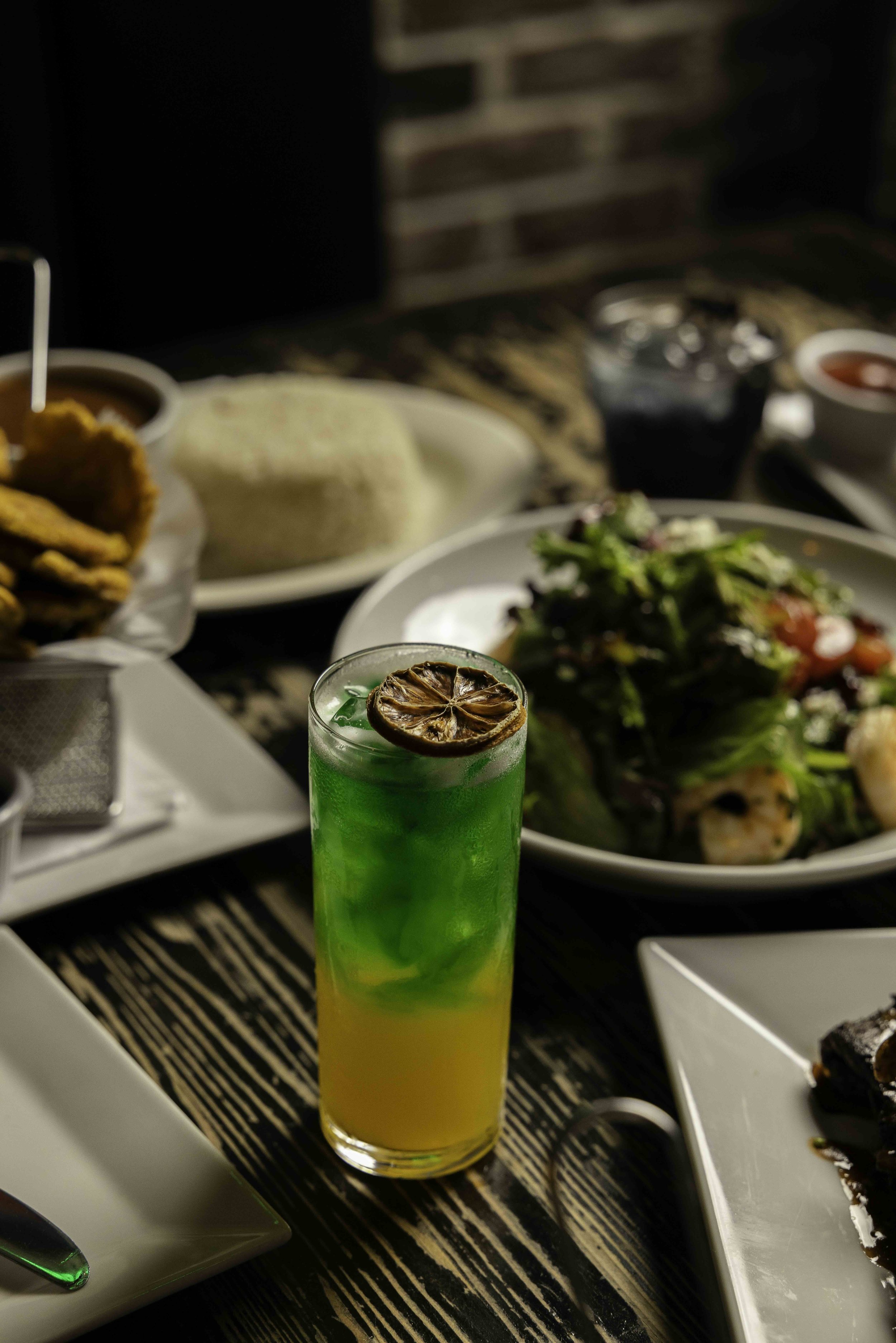 A layered colorful cocktail with a dried lemon slice garnish on top, surrounded by plates of salad, bread, fried foods, and drinks on a dark wooden table.