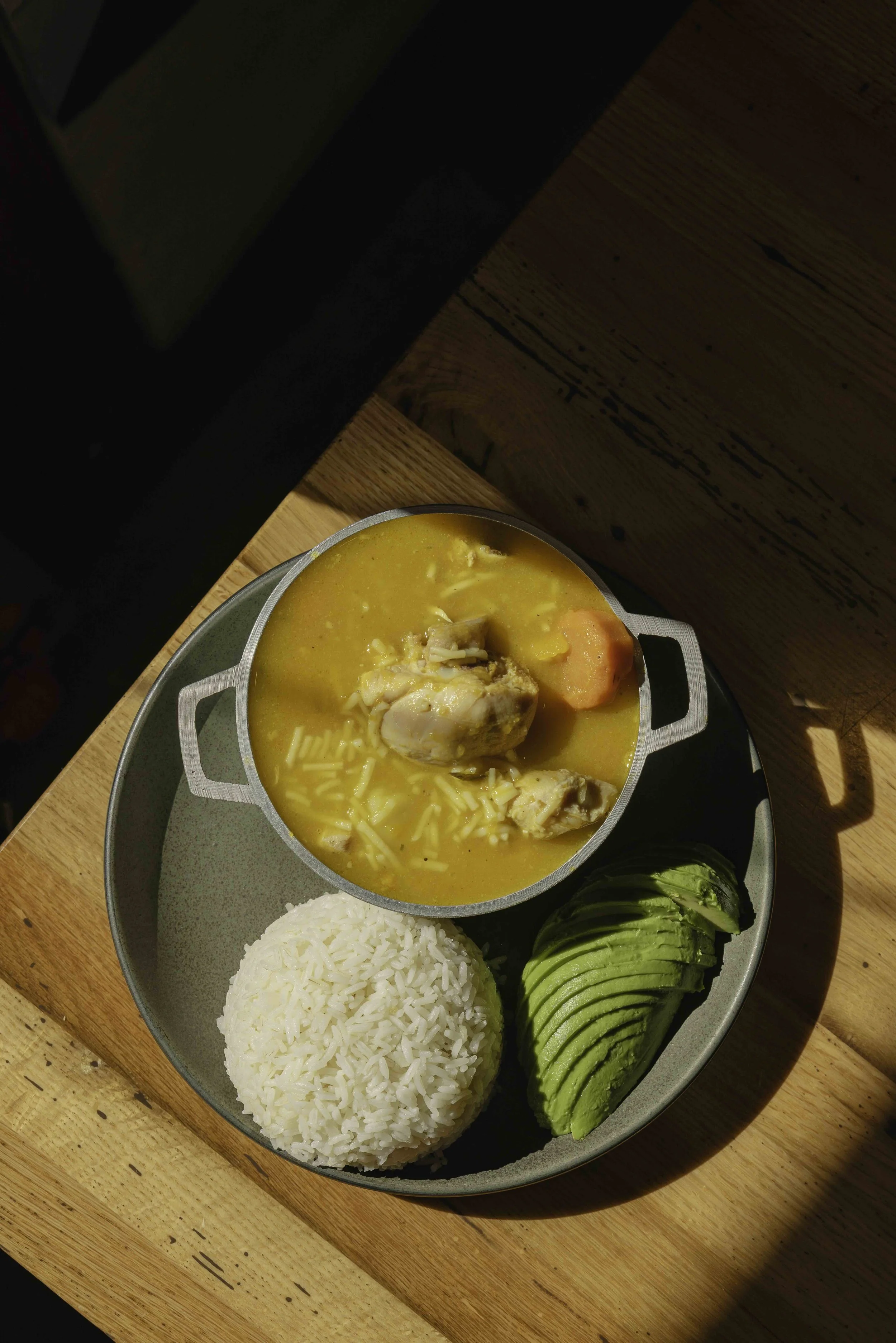 A plate with rice, sliced avocado, and a bowl of chicken and vegetable soup on a wooden table.
