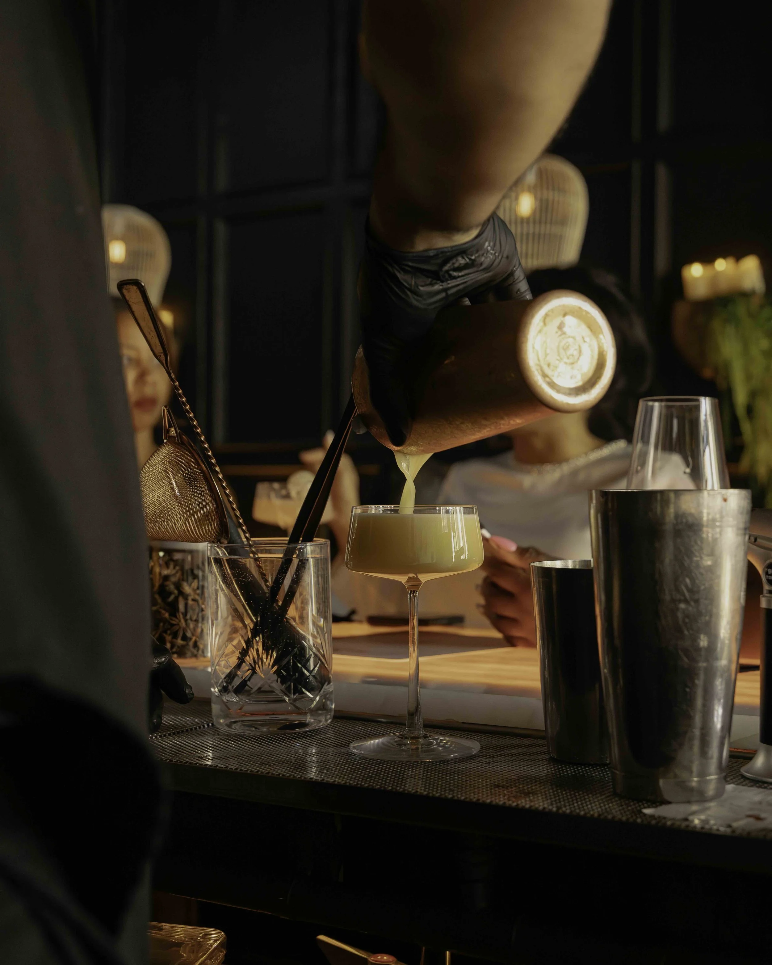 Bartender pouring a yellow cocktail from a shaker into a coupe glass at a bar.
