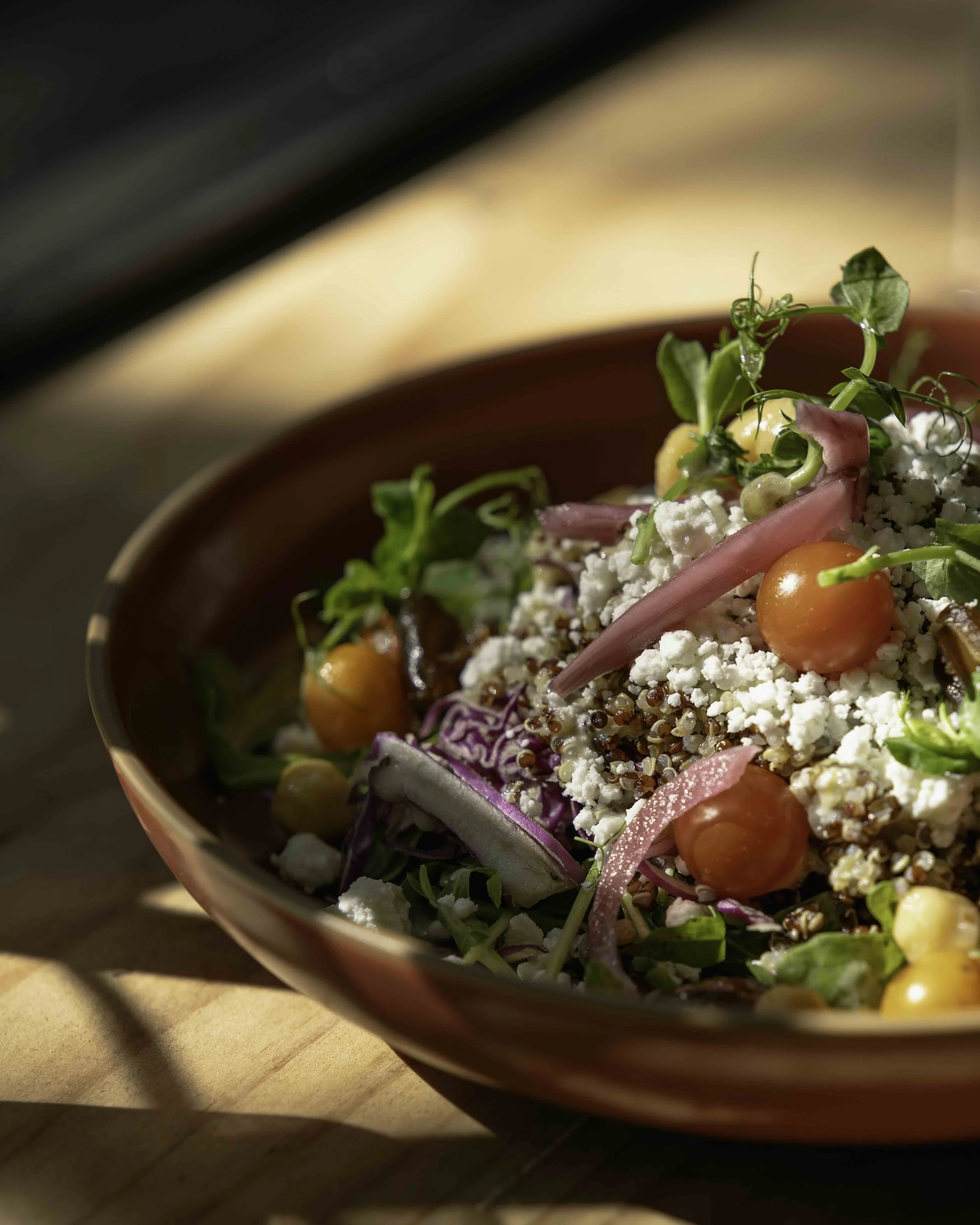 A close-up of a colorful vegetable and grain salad in a brown bowl, featuring cherry tomatoes, red onions, leafy greens, shredded cheese, quinoa, and microgreens.