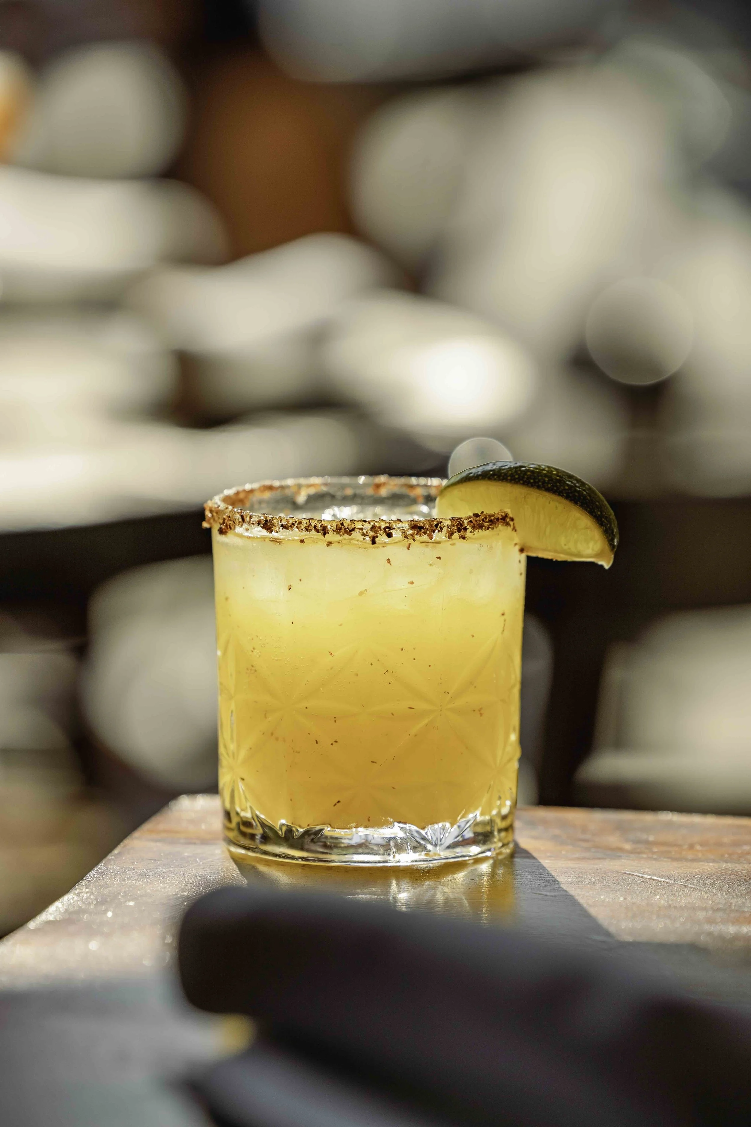 Close-up of a cocktail garnished with a lime wedge on a wooden surface, with a salted rim and a blurred background.