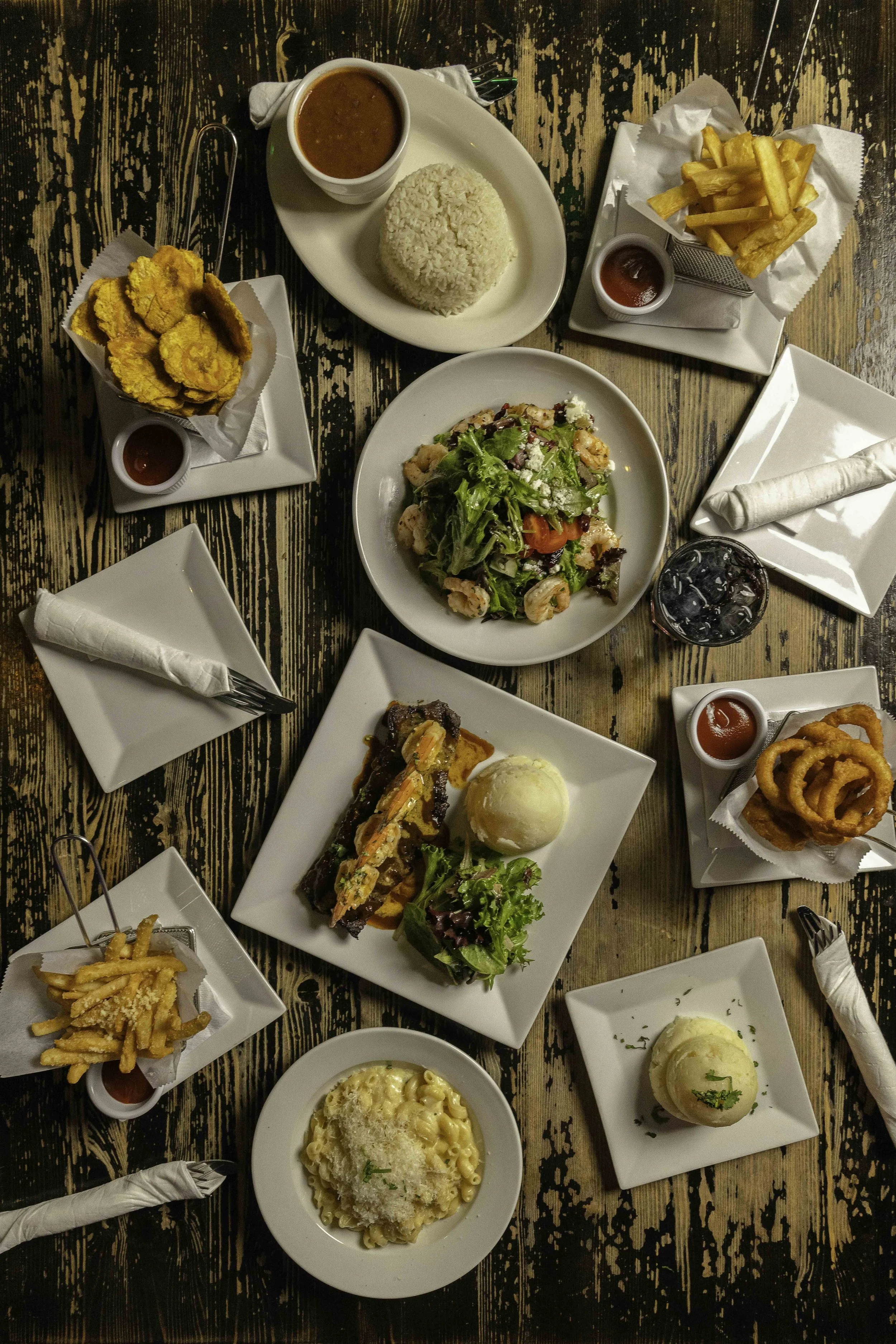 A variety of dishes on a wooden table, including fried shrimp, rice, curry, french fries, onion rings, salad, baked macaroni, and a steak with mashed potatoes, with utensils and napkins around.