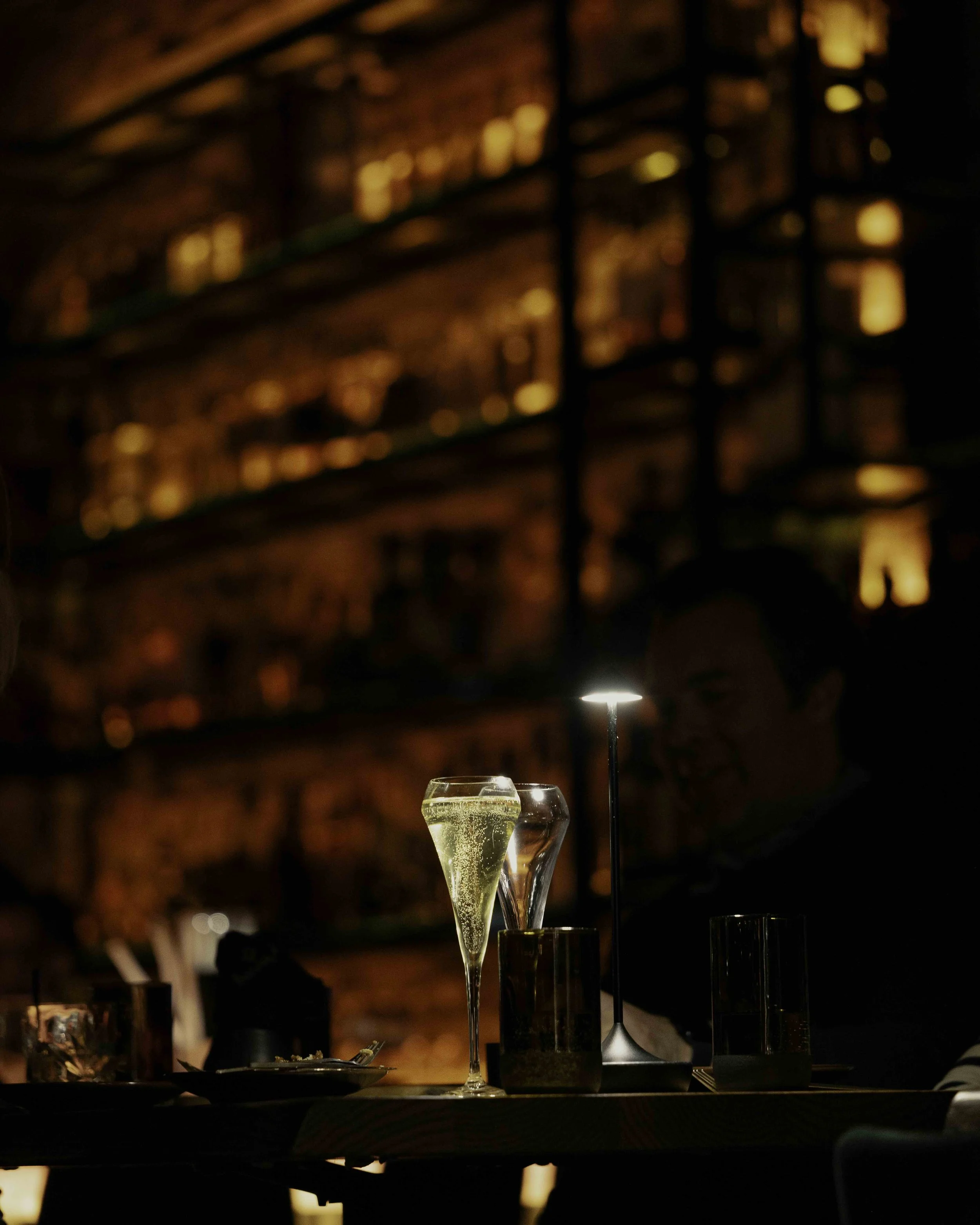A bar scene with two glasses, one filled with champagne and the other with a dark drink, on a table. A person smiling in the background, blurred, with a brick wall with shelves of bottles illuminated behind.