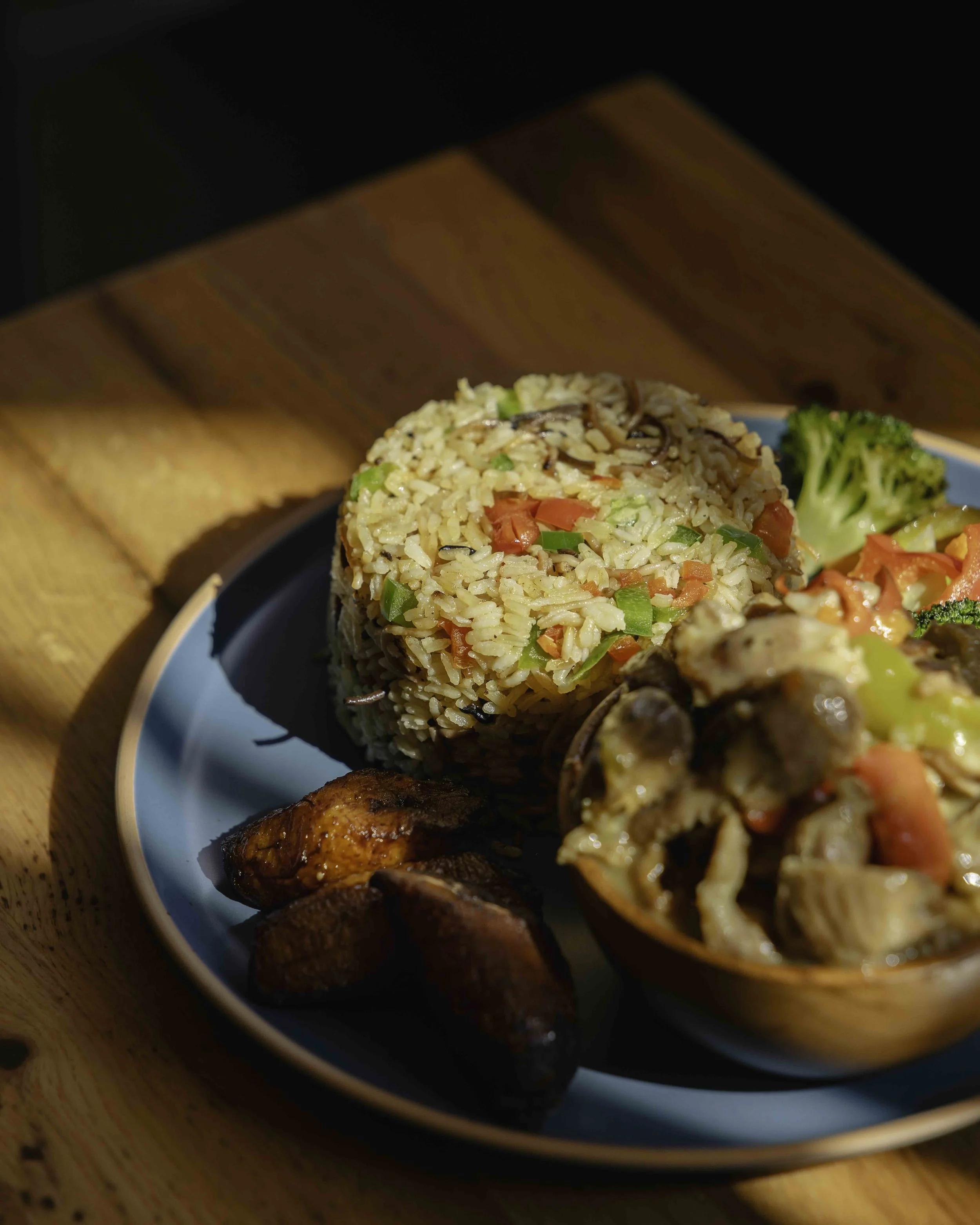 A plate of fried rice with vegetables, broccoli, a side of roasted plantains, and sautéed mushrooms and vegetables.