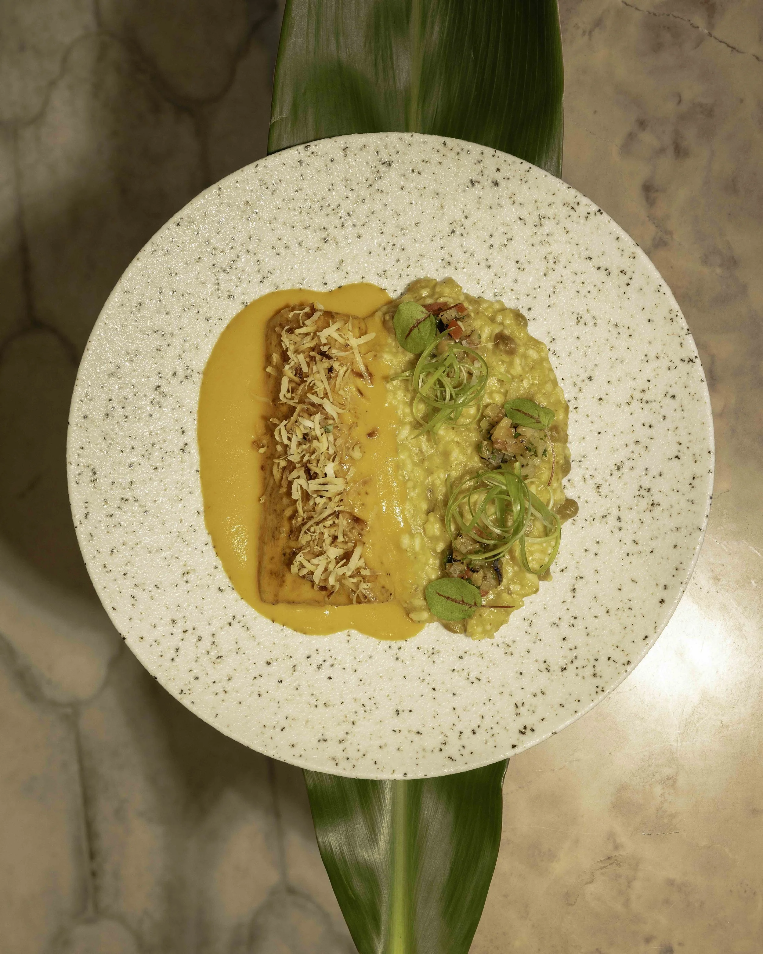 Plate of risotto garnished with microgreens and red pepper flakes, served with a side of shredded cheese and topped with yellow sauce, on a white speckled plate with a green leaf base.