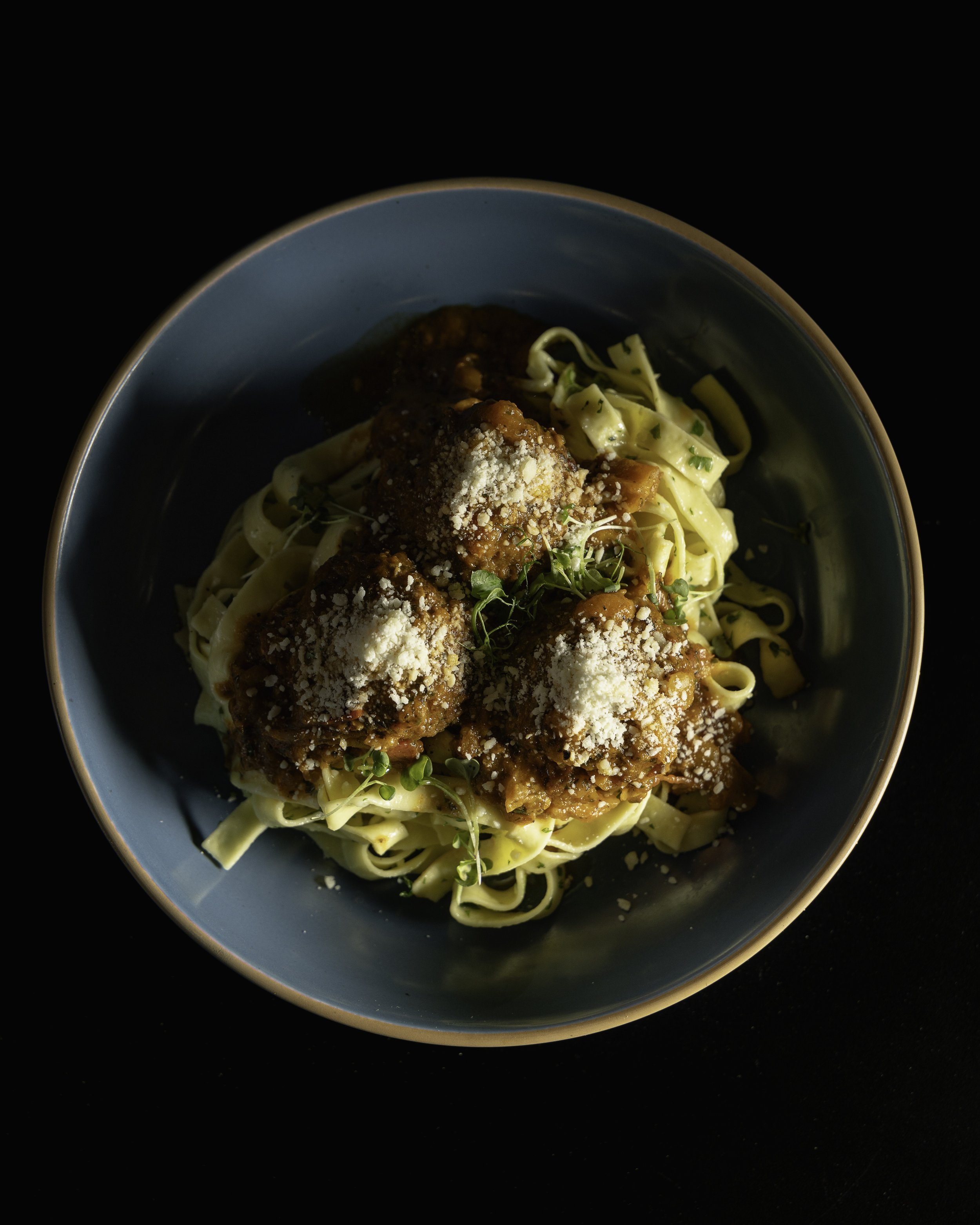 Plate of pasta topped with meatballs, tomato sauce, grated cheese, and herbs, in a blue bowl on a dark surface.
