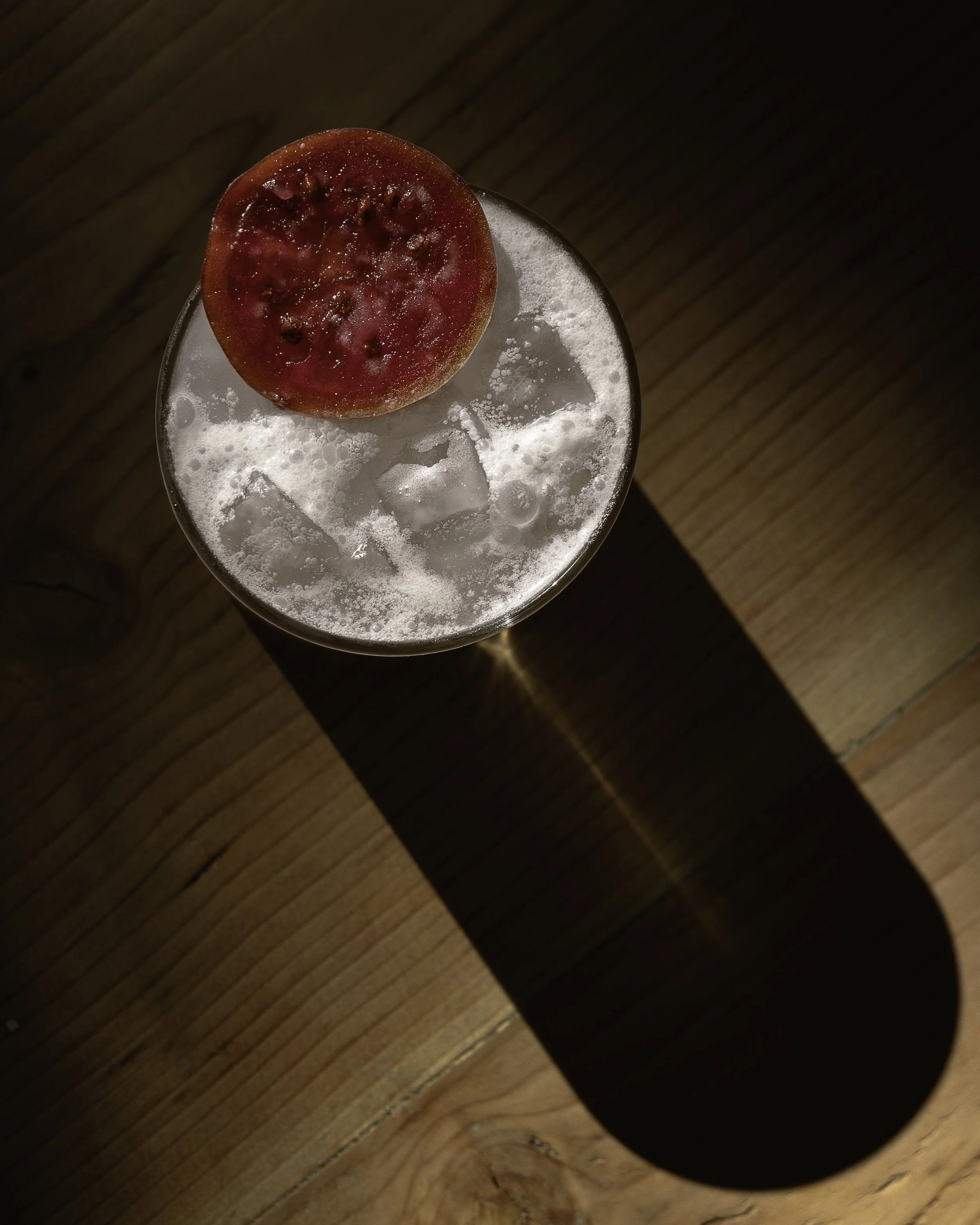 A glass of clear bubbly drink with ice cubes, garnished with a halved small red fruit, possibly a small apple or plum, on a wooden surface with a long shadow cast by the glass.