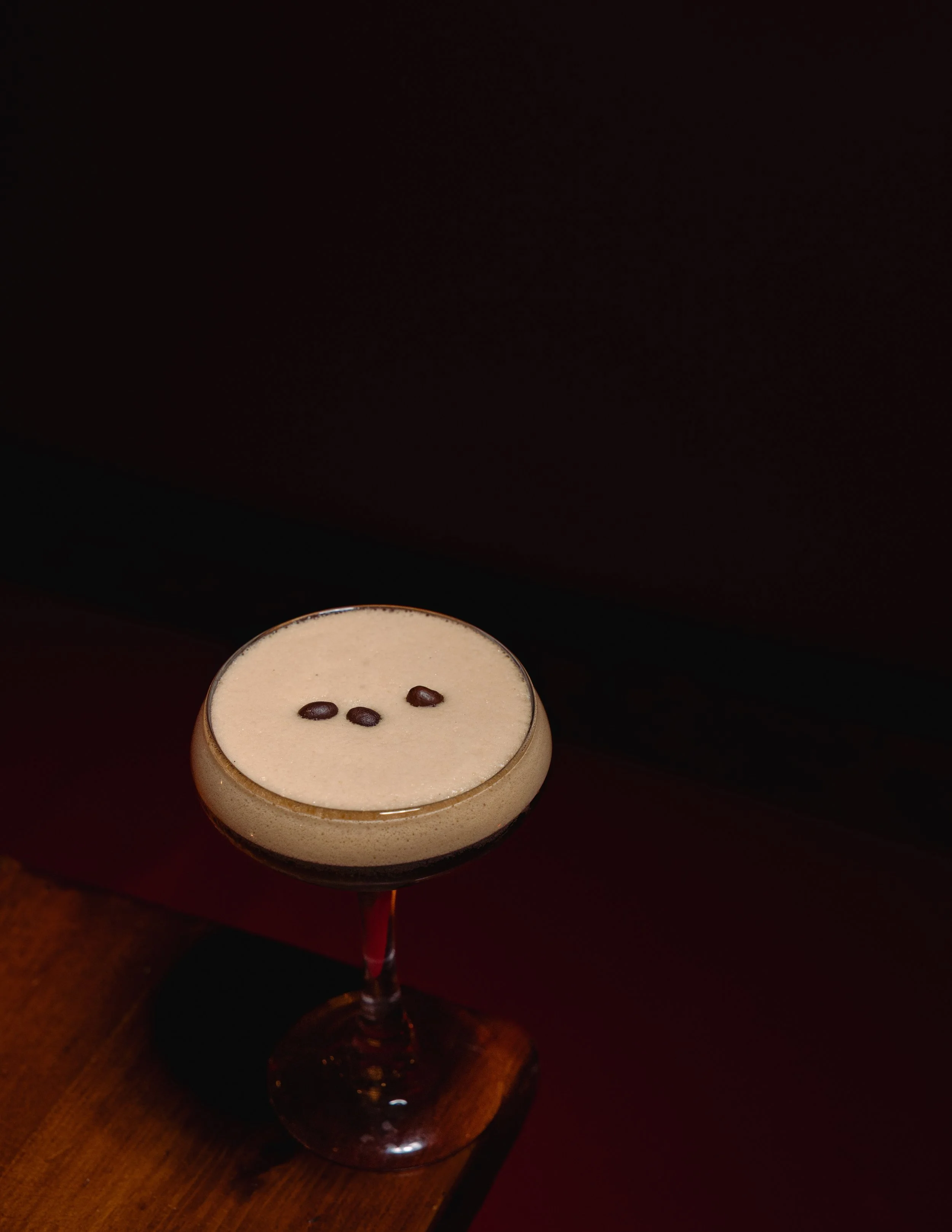 A cocktail in a stemmed glass with a frothy top and three coffee beans on it, placed on a wooden surface against a dark background.