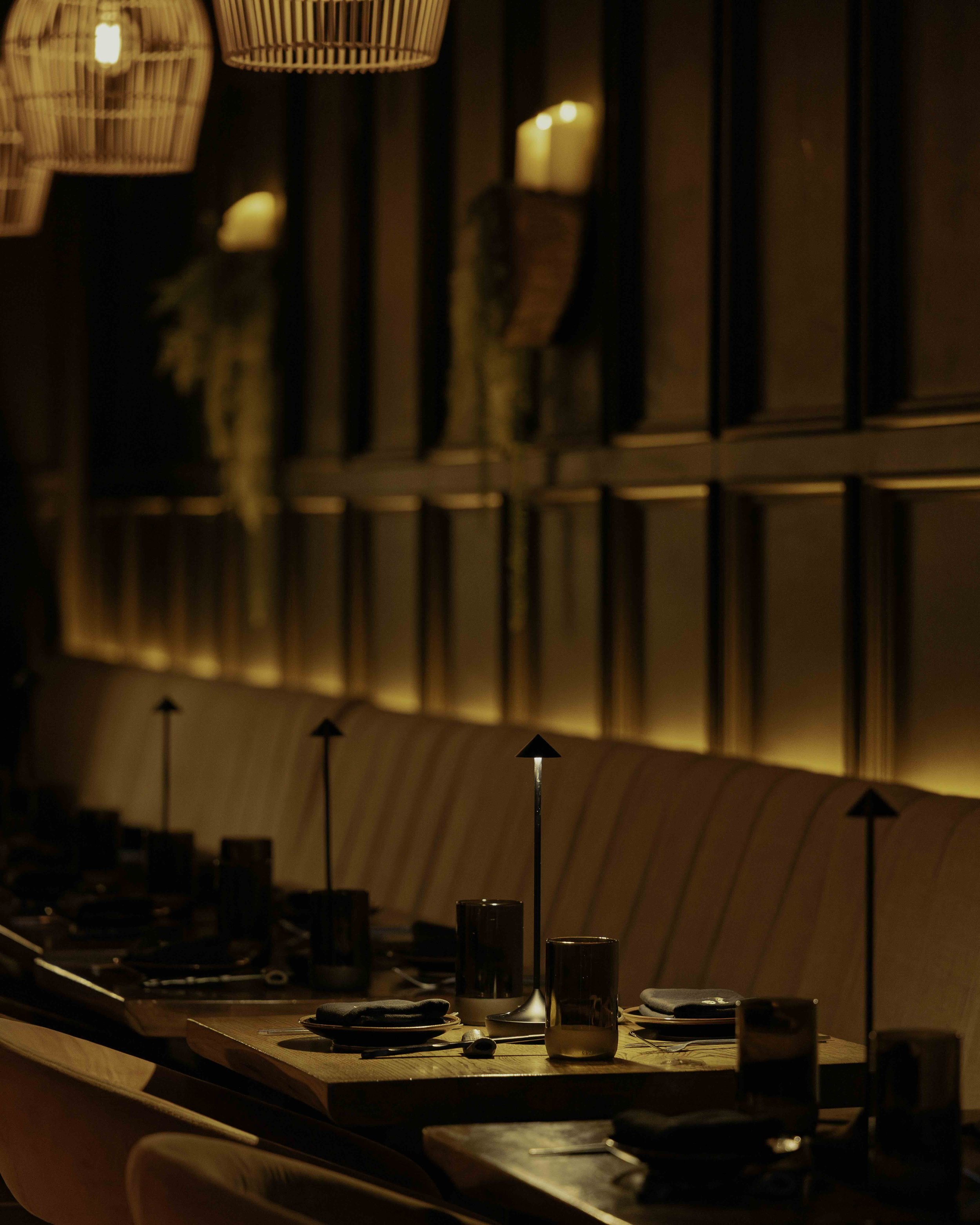 A warmly lit restaurant or dining room with a long wooden table set with glasses, plates, and black napkins, surrounded by cushioned seating and decorative wooden wall panels.