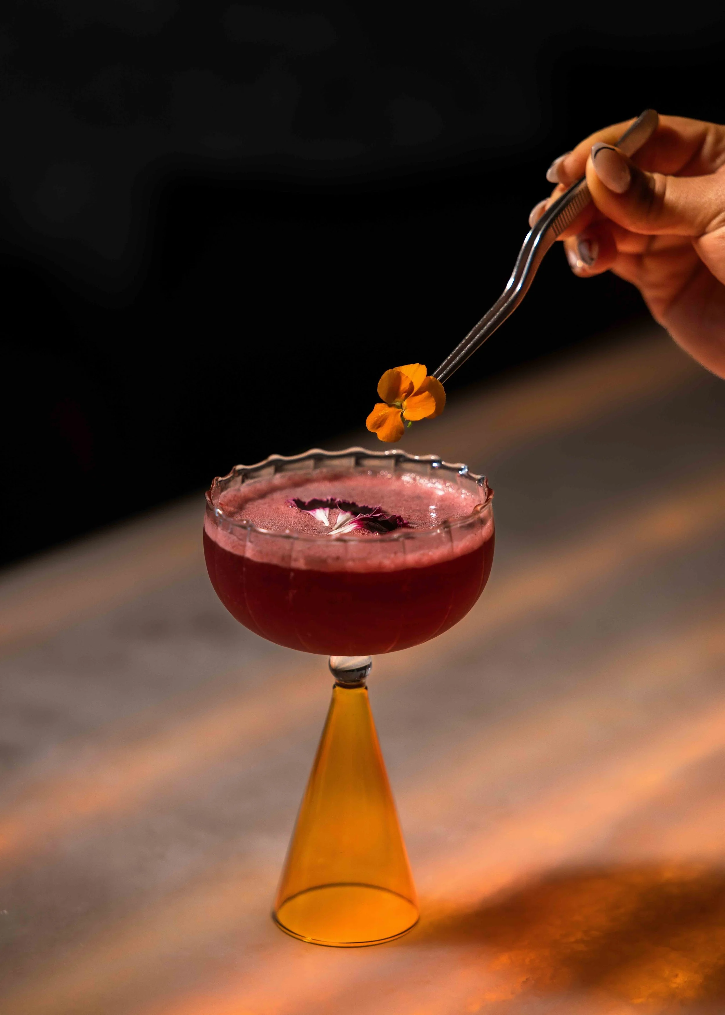 A person's hand using tongs to place an orange edible flower on top of a red cocktail in a vintage glass with a gold bottom, set against a dark background.