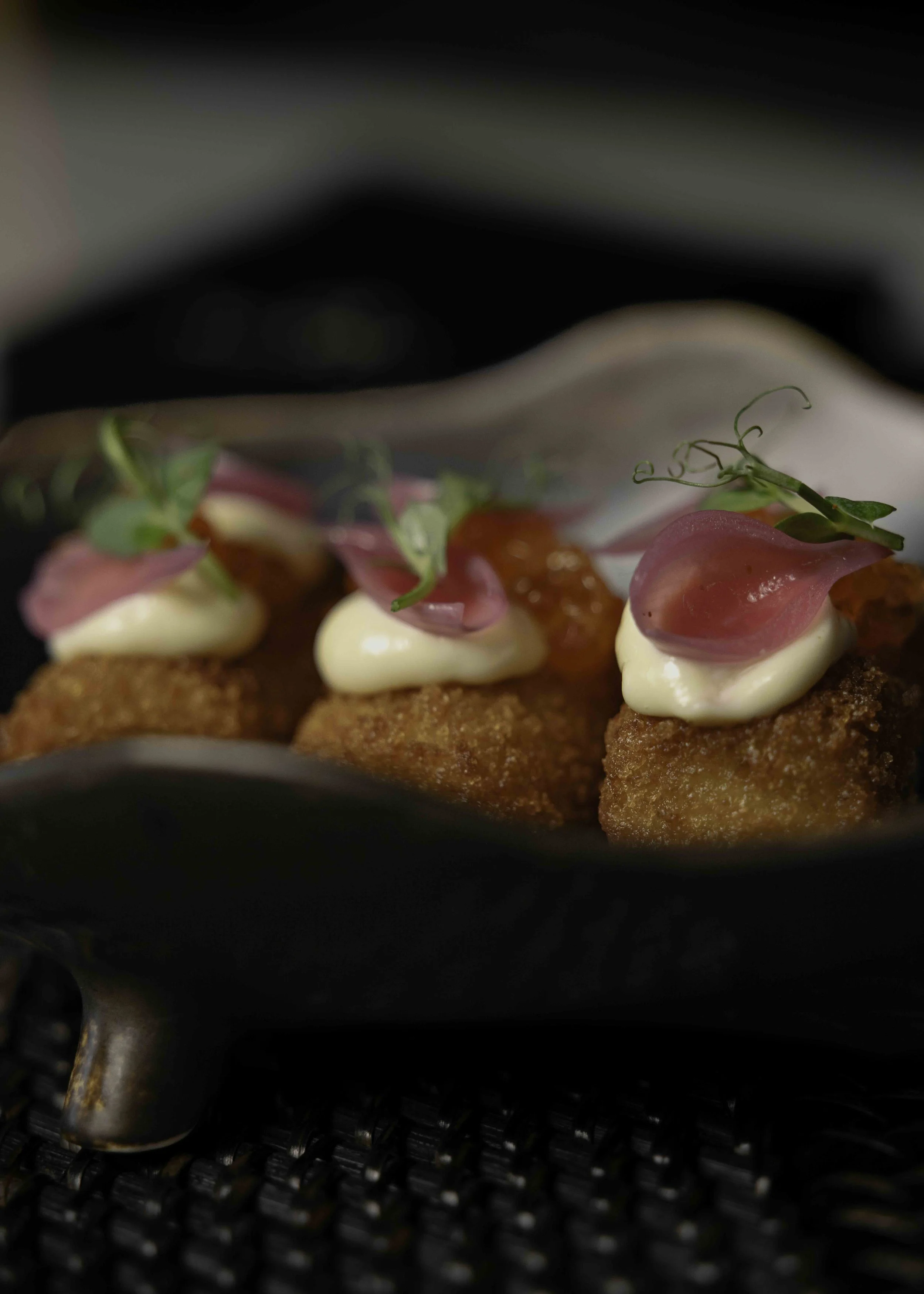 Close-up of three breaded fried appetizers topped with dollops of white sauce, pink pickled onions, and microgreens on a black plate.