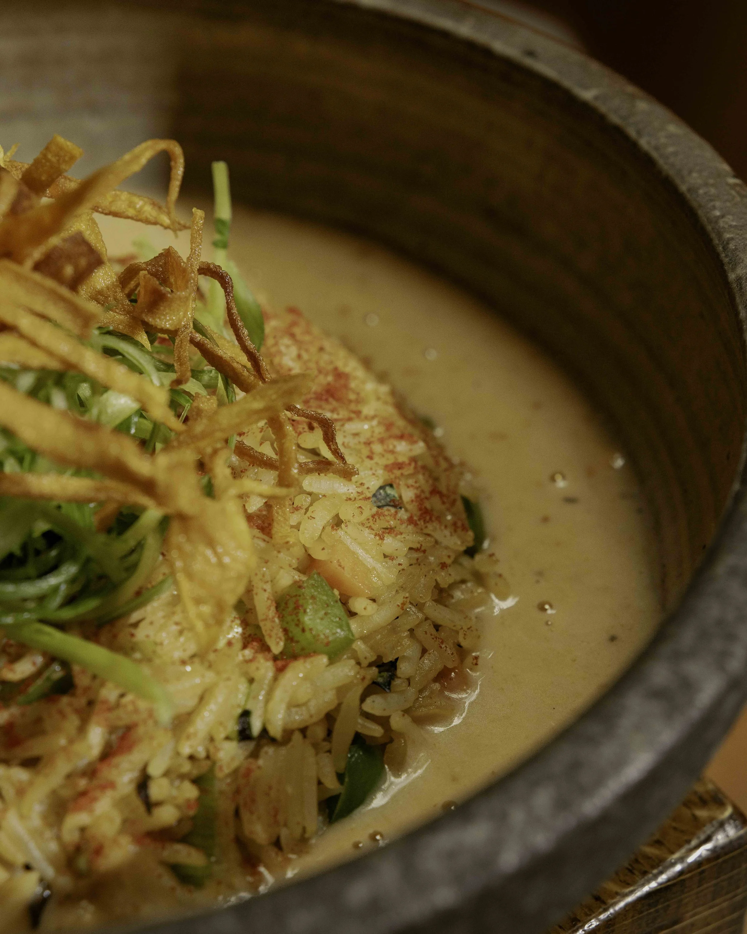 Close-up of a bowl of rice topped with crispy fried onions, chopped green onions, red paprika, and a creamy soup or sauce around the rice inside a dark bowl.
