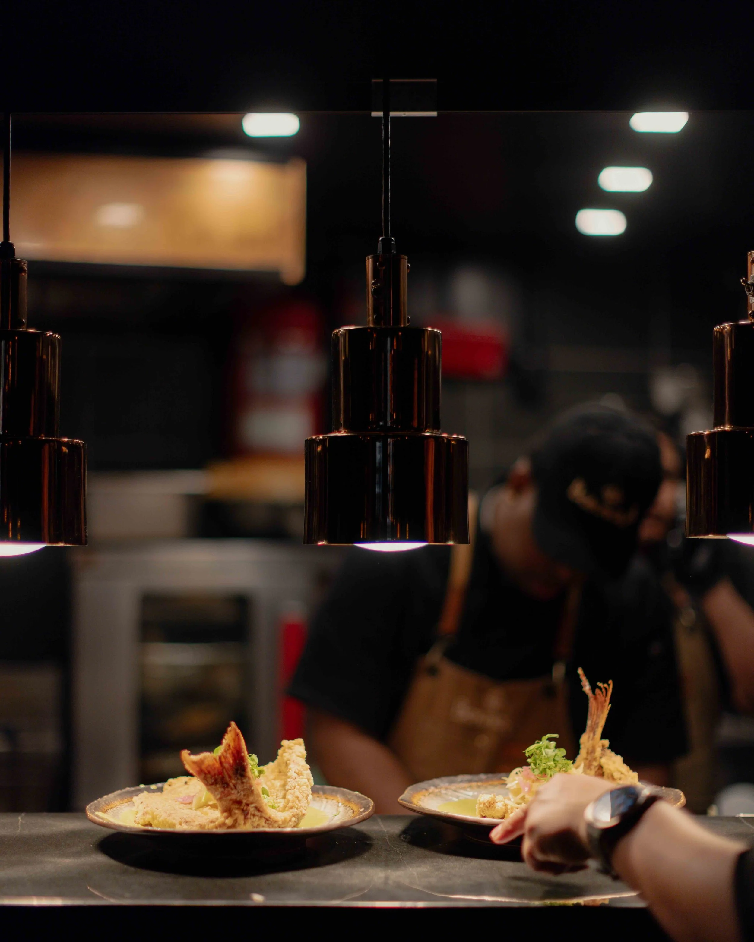 Two plates of tempura prawns and vegetables, garnished with green herbs, on a black counter in a dark, modern restaurant kitchen.