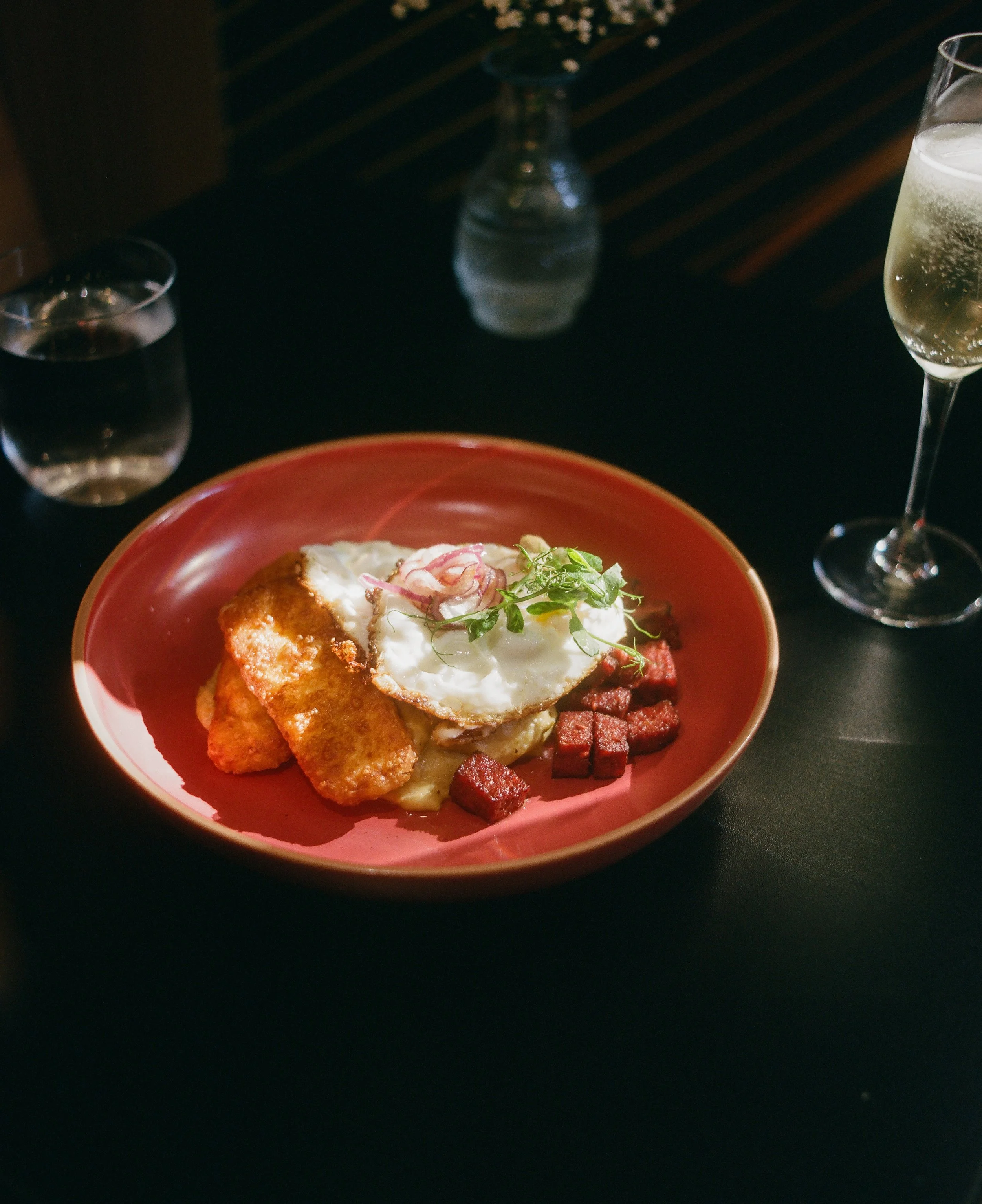 A plate of breakfast with fried fish, a fried egg, crispy potato wedges, and diced caramelized ham, garnished with a sprig of microgreens, on a dark table with a water glass, a glass of champagne, and a small vase with white flowers in the background