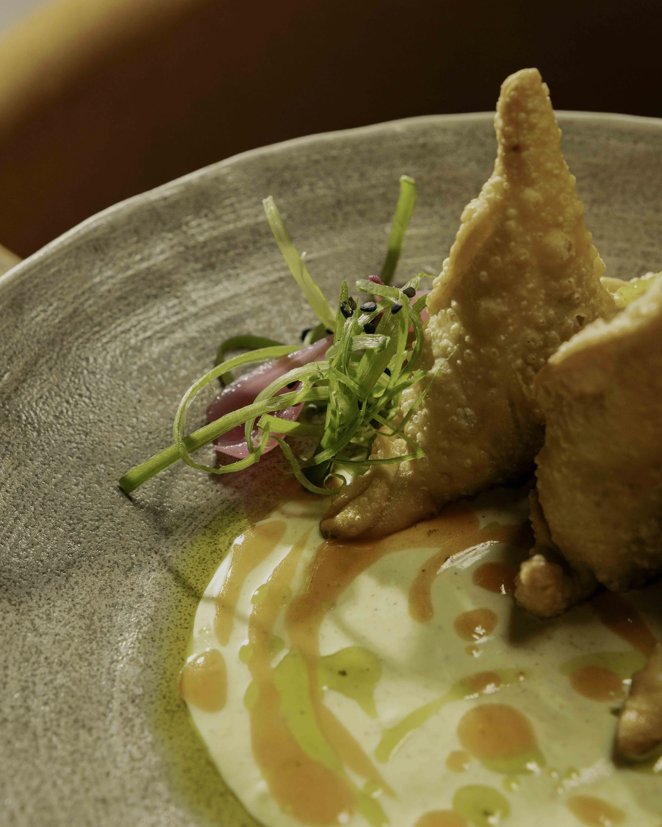 Close-up of a plated dish with two crispy fried pieces, garnish of microgreens and sliced radish, drizzled with sauces on a textured beige plate.