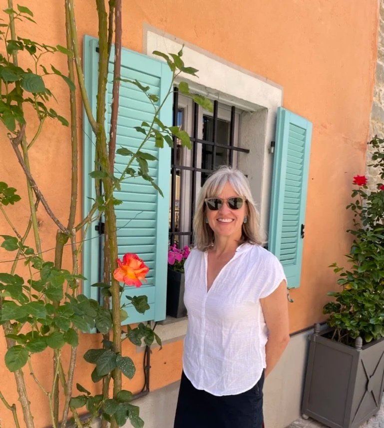 Woman with blonde hair and sunglasses smiling outside a house with turquoise shutters, a orange wall, and potted flowers.