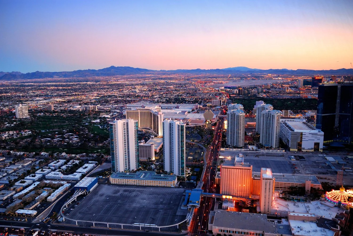 Aerial view of a city at dusk with tall buildings, streets illuminated, mountains in the background, and a colorful sky.