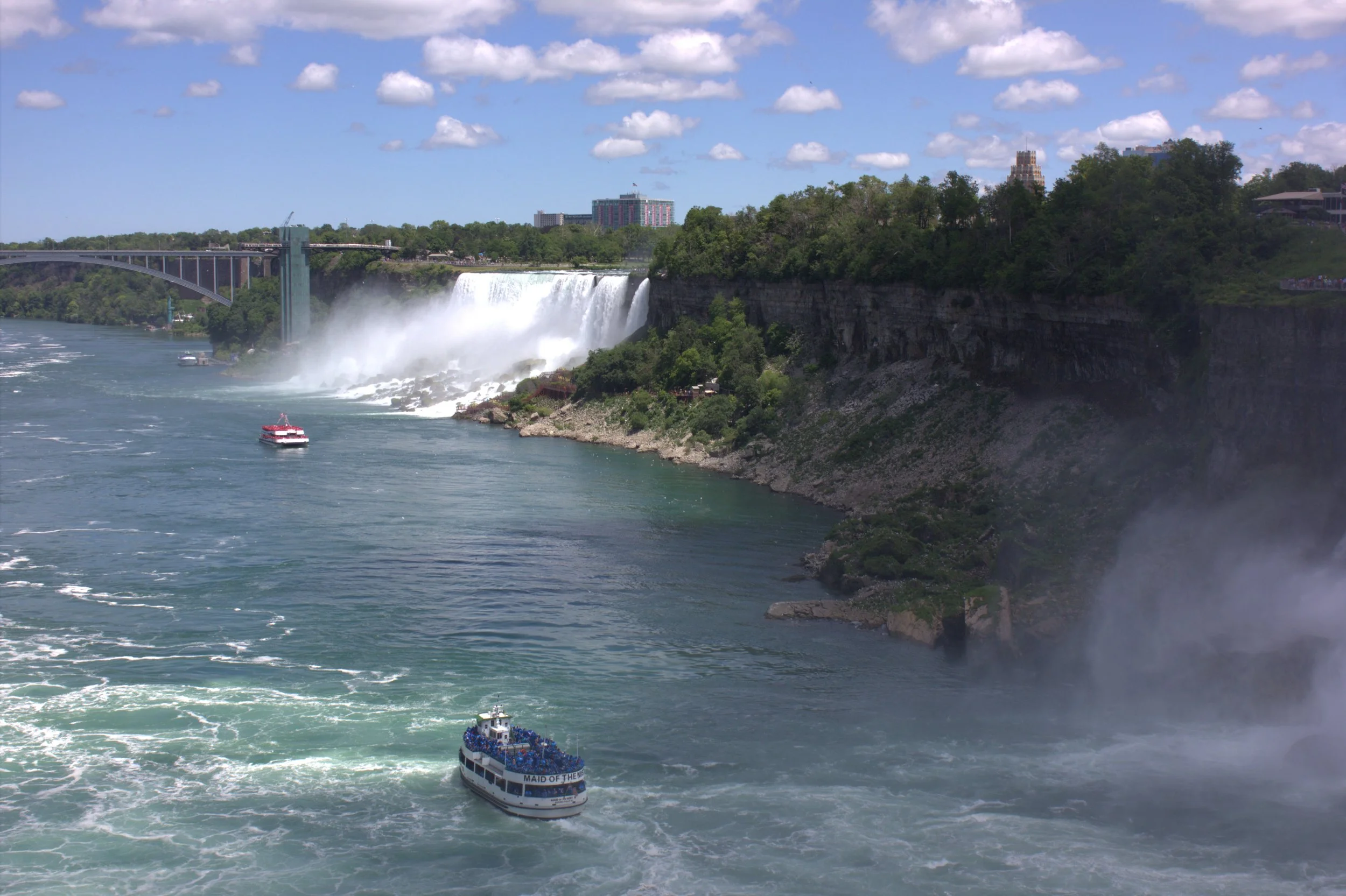 View of Niagara Falls with boats on the water and a bridge in the background under a partly cloudy sky.