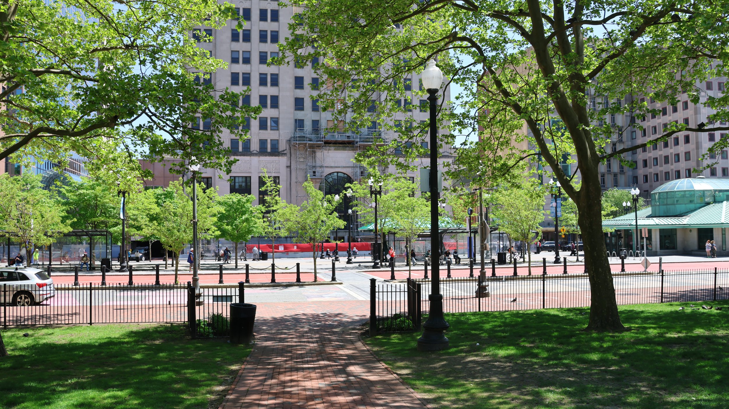 Urban park viewed through trees with benches, streetlamps, a bus stop, and tall buildings in the background on a sunny day.
