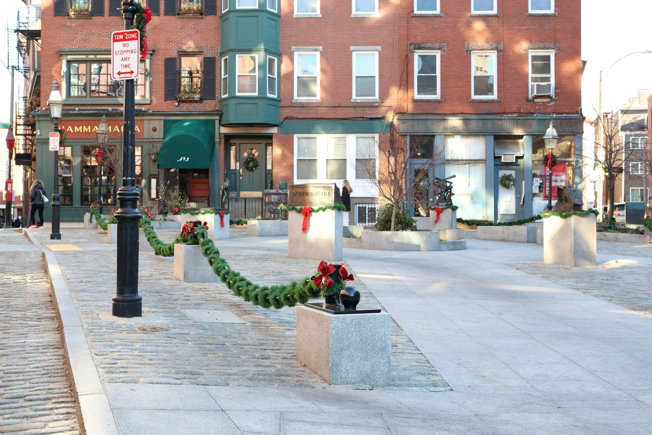 A city street scene decorated for the holidays with green garlands, red ribbons, and holiday wreaths. There are flower planters with festive bows, and people are walking by. The buildings are brick and have large windows, some with window air conditioning units. There are street signs and old-fashioned street lamps.