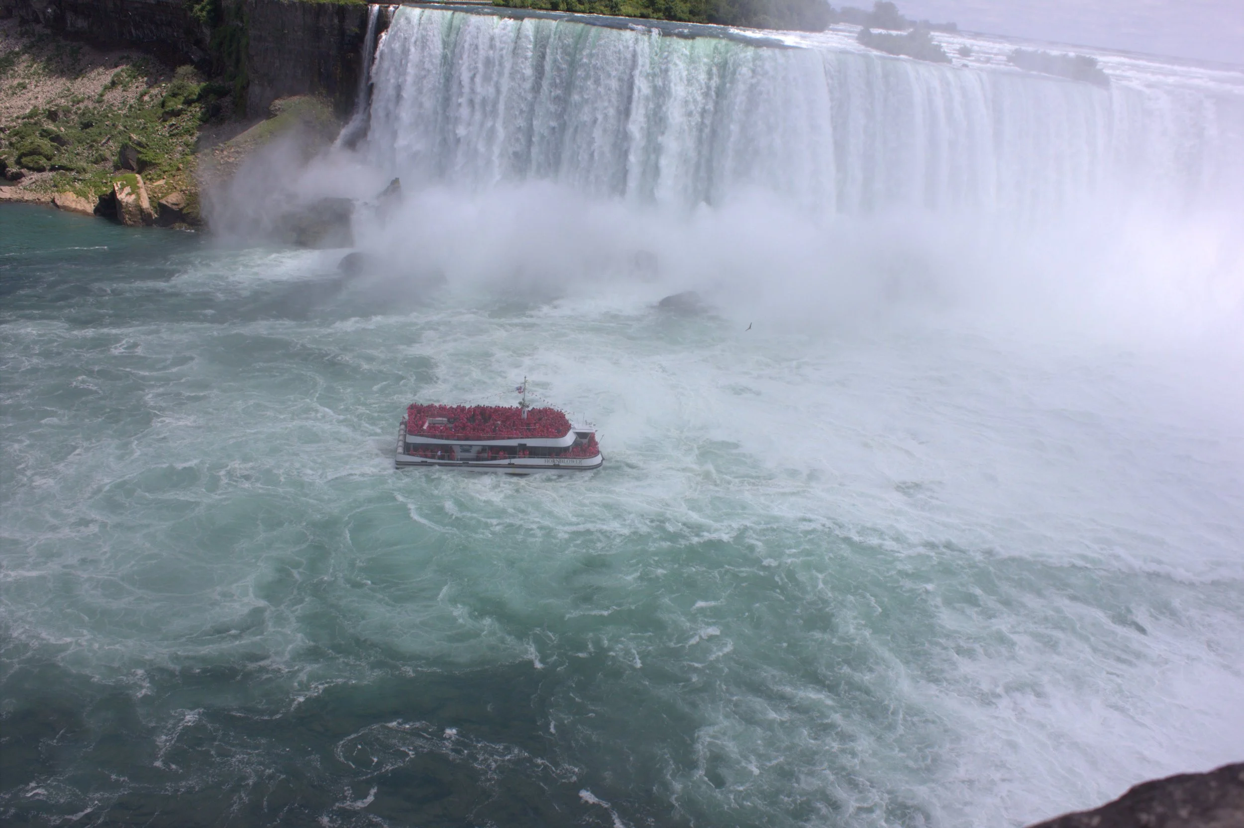 A view of Niagara Falls with a boat near the waterfall, surrounded by mist and green landscape.