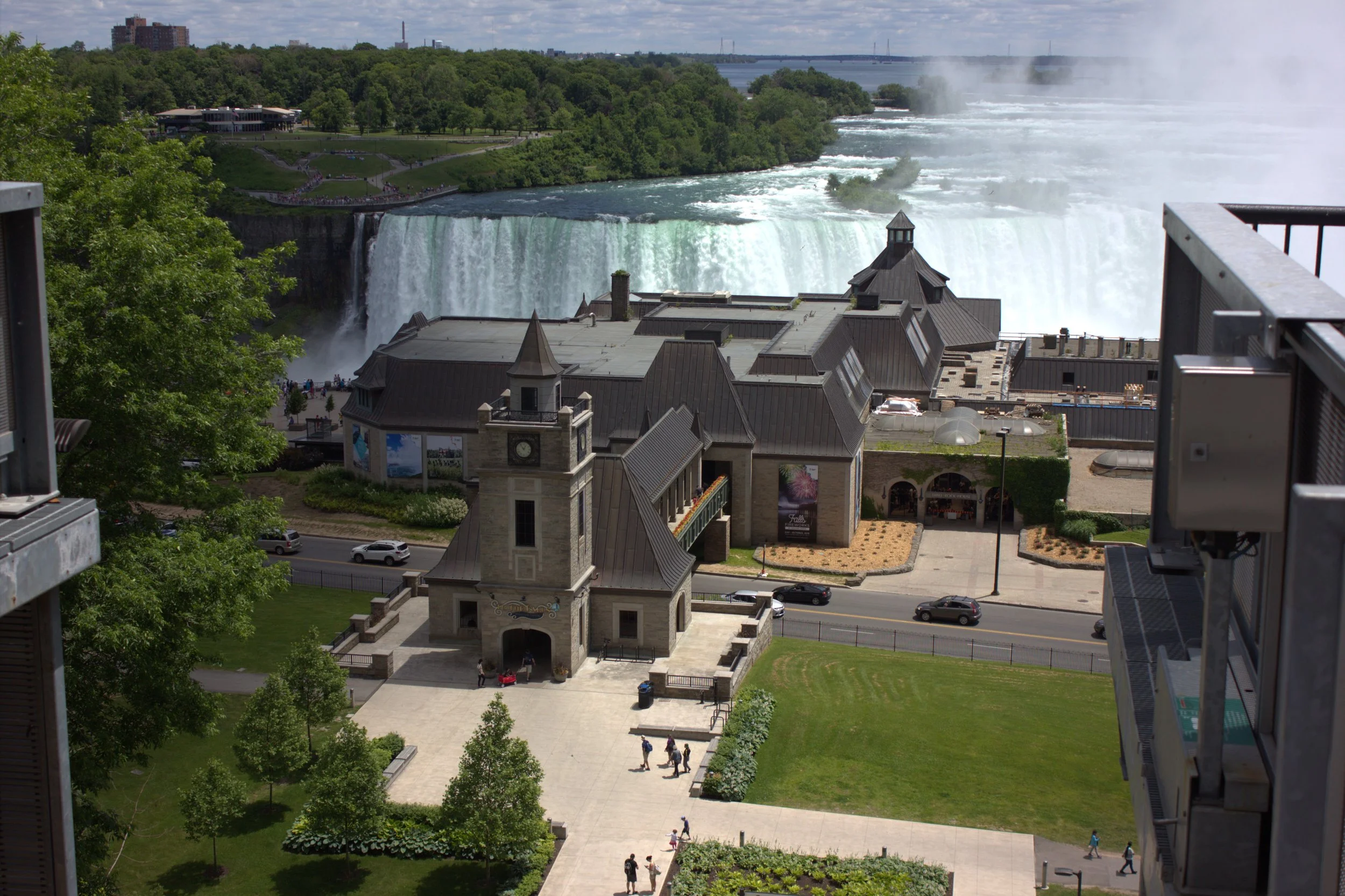View of Niagara Falls with a building in the foreground, a park with a walking path, and trees, with the river and bridge in the background.