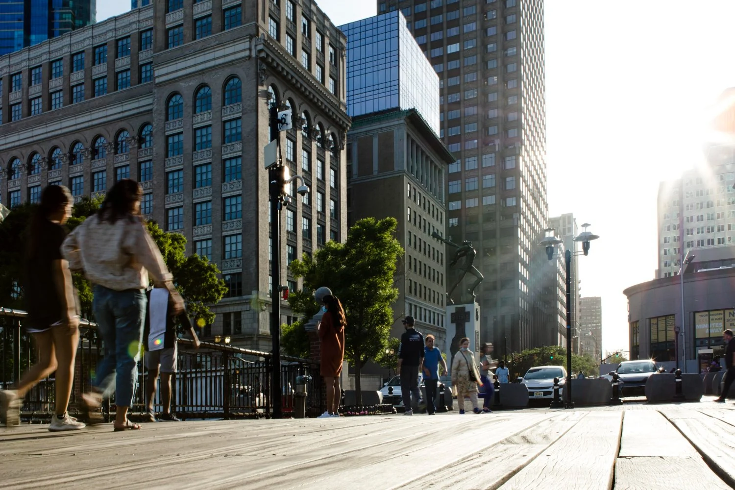 People walking on a city sidewalk with tall buildings and storefronts in the background, sunlight shining from the right.