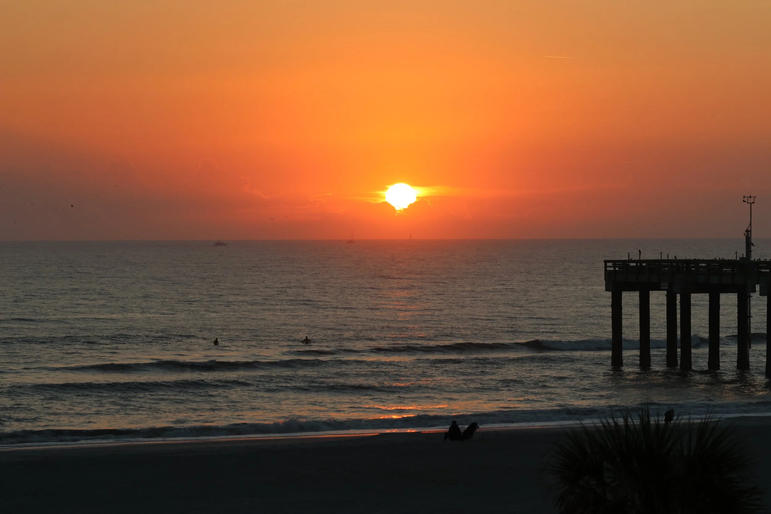 Sunset over the ocean with a pier extending into the water, silhouettes of people sitting on the beach, and boats visible in the distance.