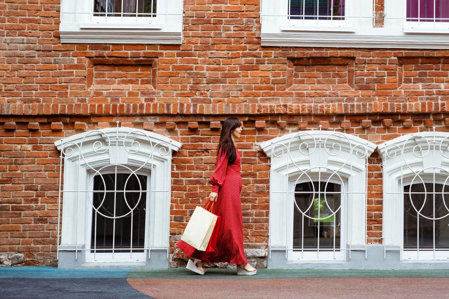 A woman wearing a long red dress and white shoes walking along a brick building with white decorative window frames, carrying shopping bags.