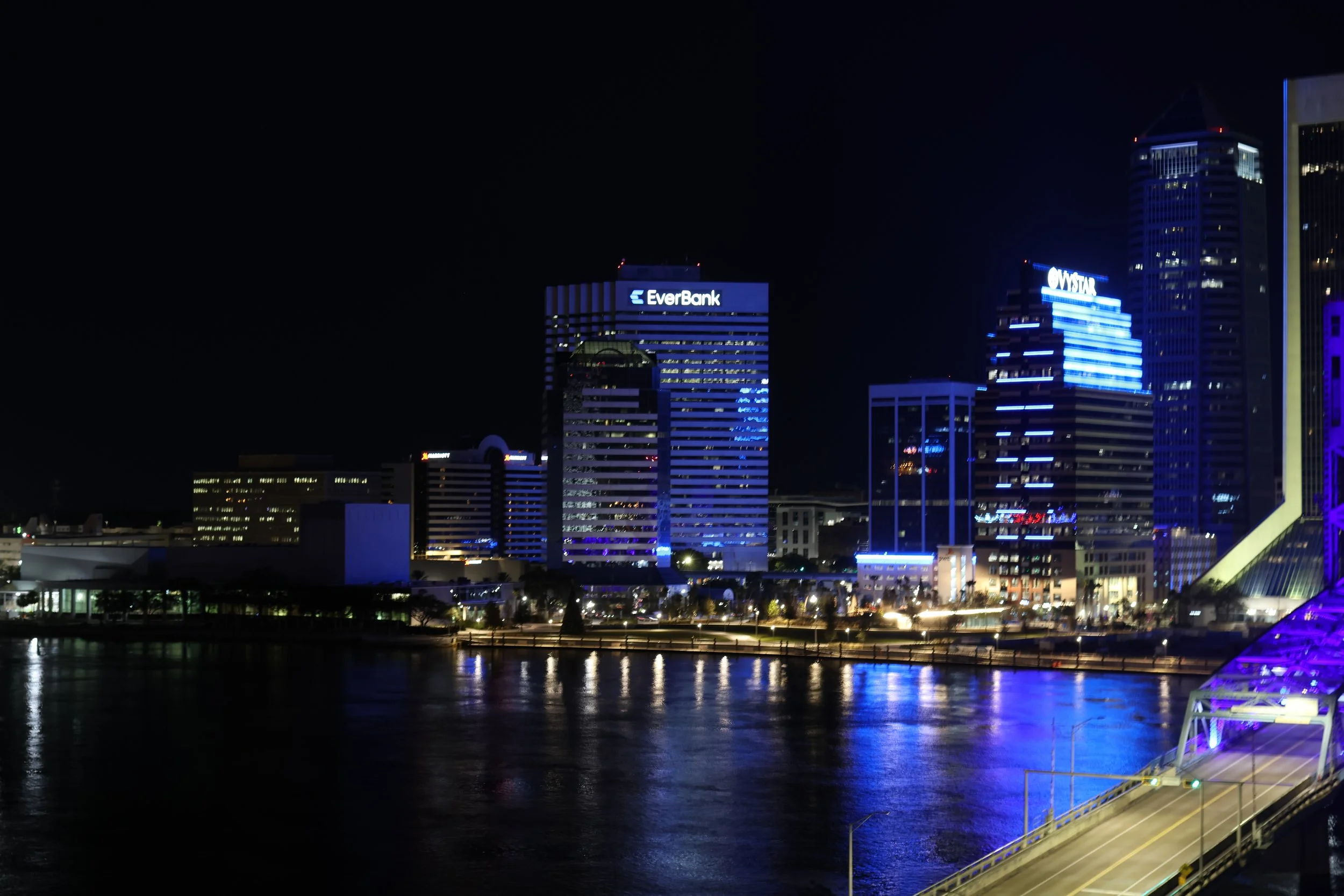 Night skyline of a city with tall buildings illuminated in blue and white lights, reflecting on a water body in the foreground, and a waterfront area with streetlights and a road.