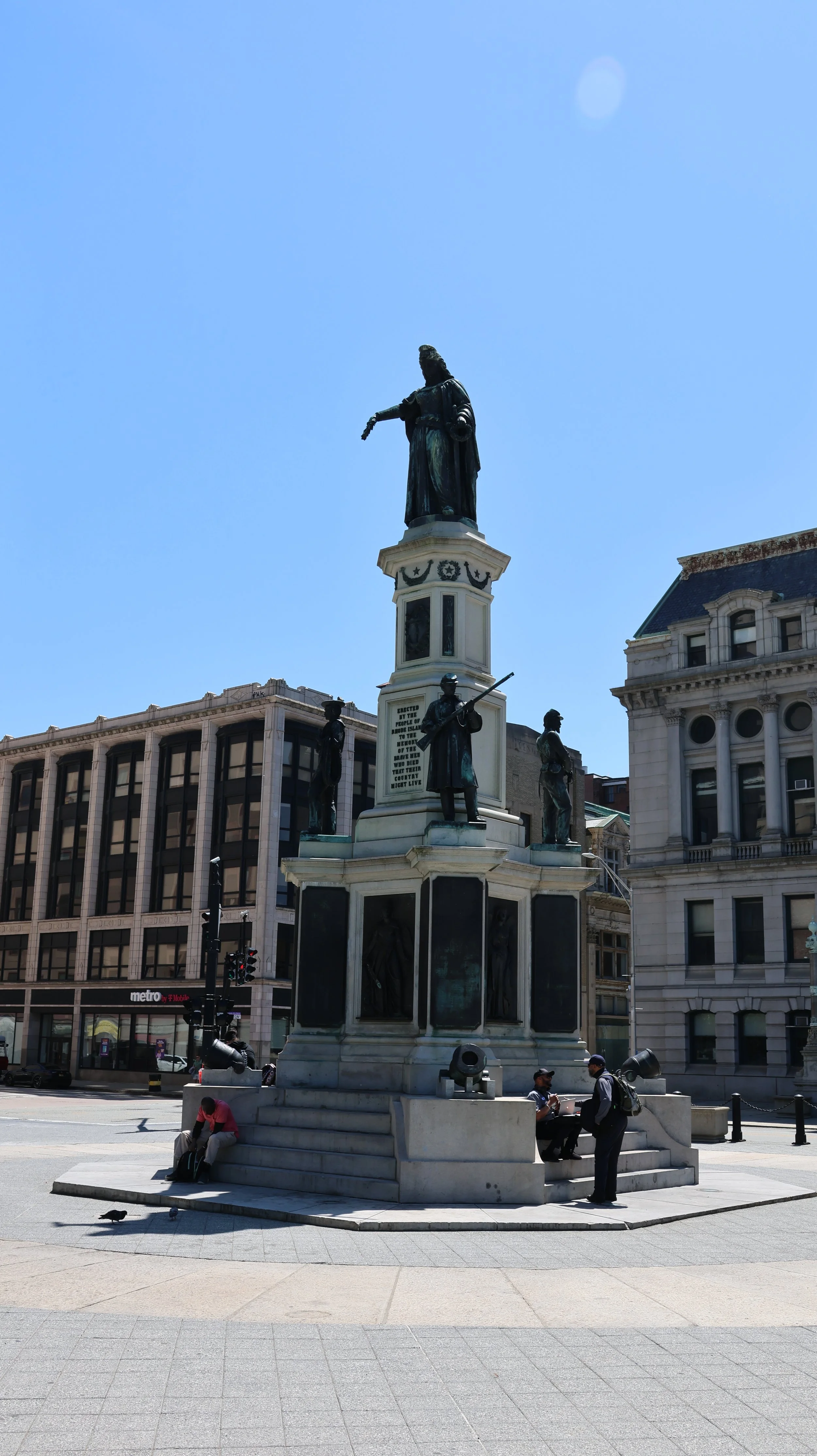 Statue monument featuring a central figure on top, surrounded by four smaller figures, in a city square with office buildings and a metro sign in the background.
