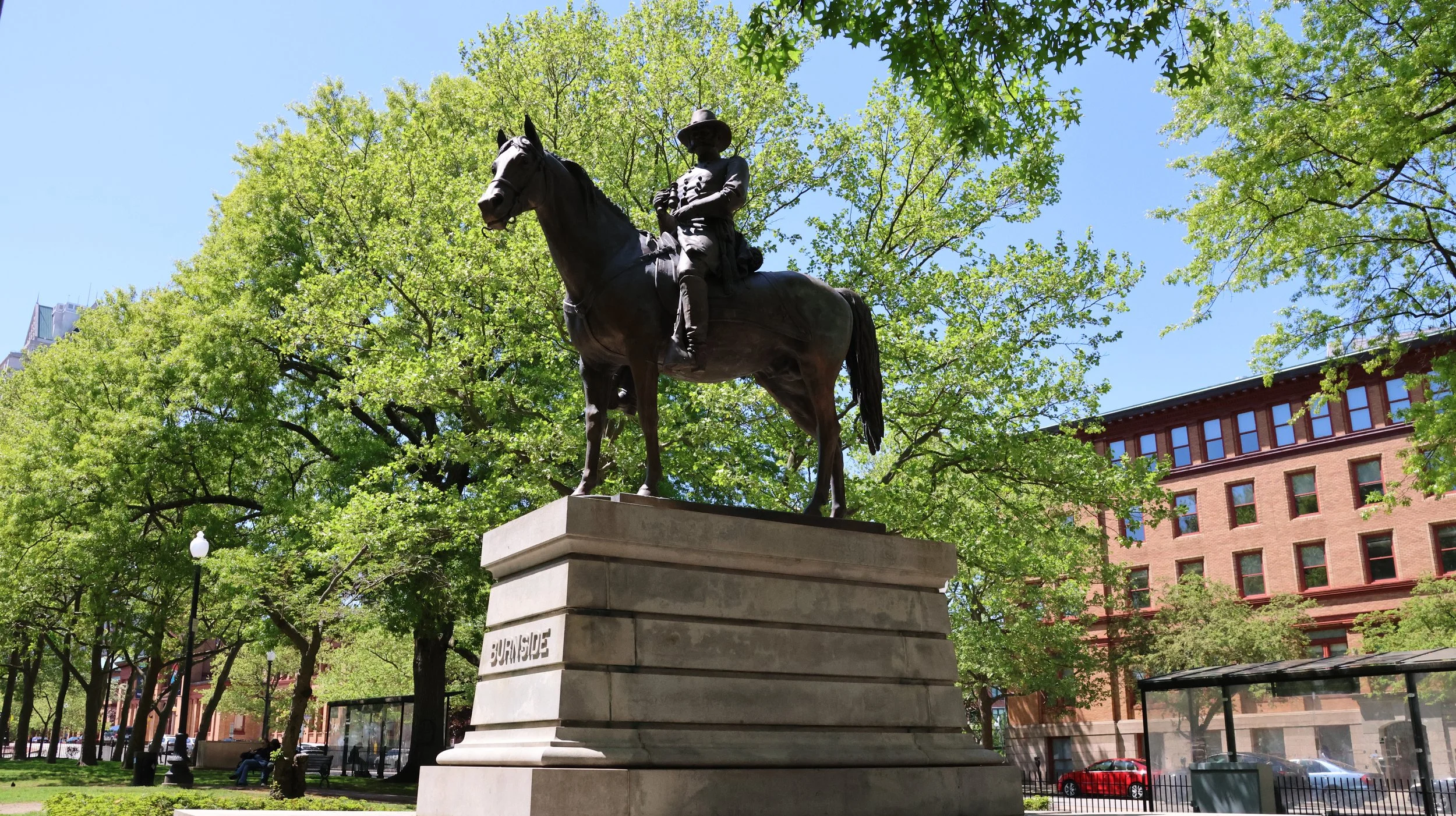 Bronze statue of a man on horseback on a stone pedestal in a park with green trees and buildings in the background.