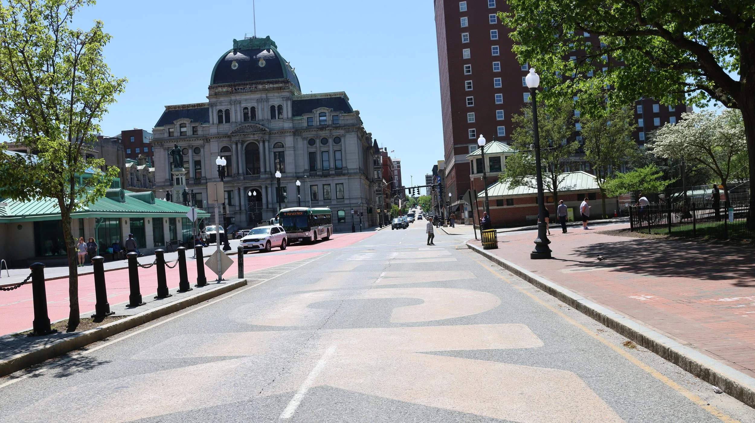 A city street scene with historic and modern buildings, trees, pedestrians, and a bus stopped at a crosswalk on a sunny day.