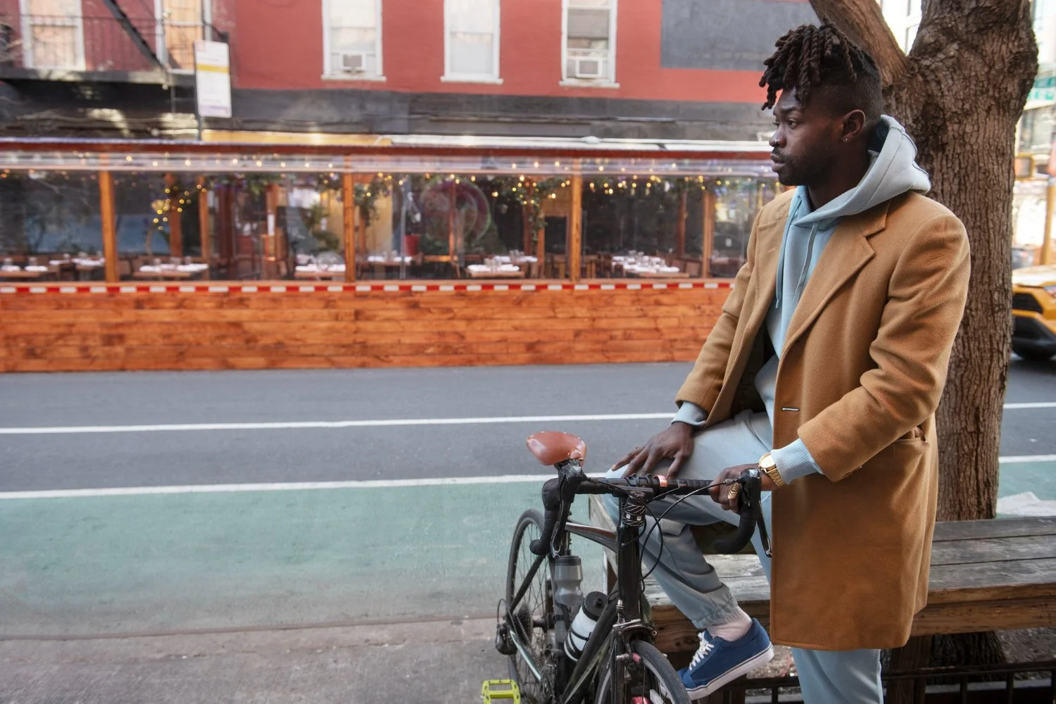 A man with dreadlocks wearing a tan coat, gray hoodie, and gray pants leaning on a black bicycle while sitting on a wooden bench under a tree on a city street. In the background, there is a restaurant with large windows and string lights inside.