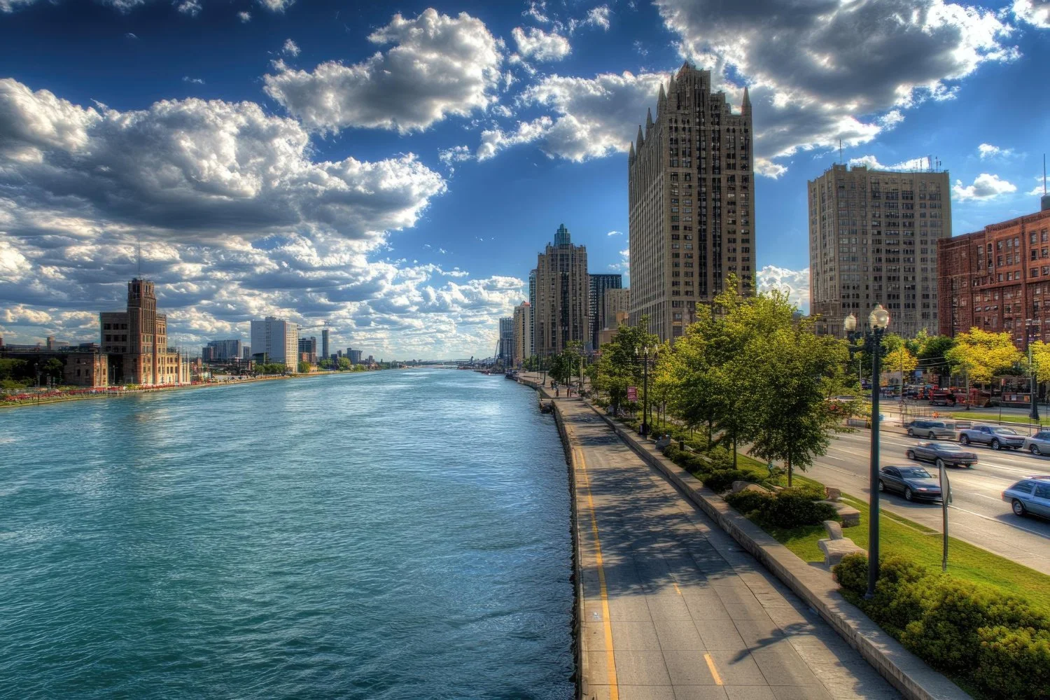 City skyline along a river with tall buildings, a blue sky with scattered clouds, a sidewalk with trees, and cars on the street.