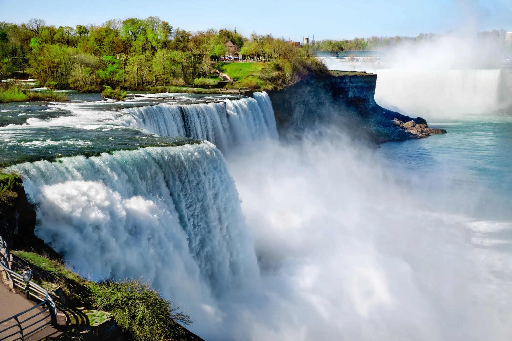 Niagara Falls with waterfalls, green trees, and observation falls in the foreground, a small pathway, and a city skyline with buildings and a bridge in the background.