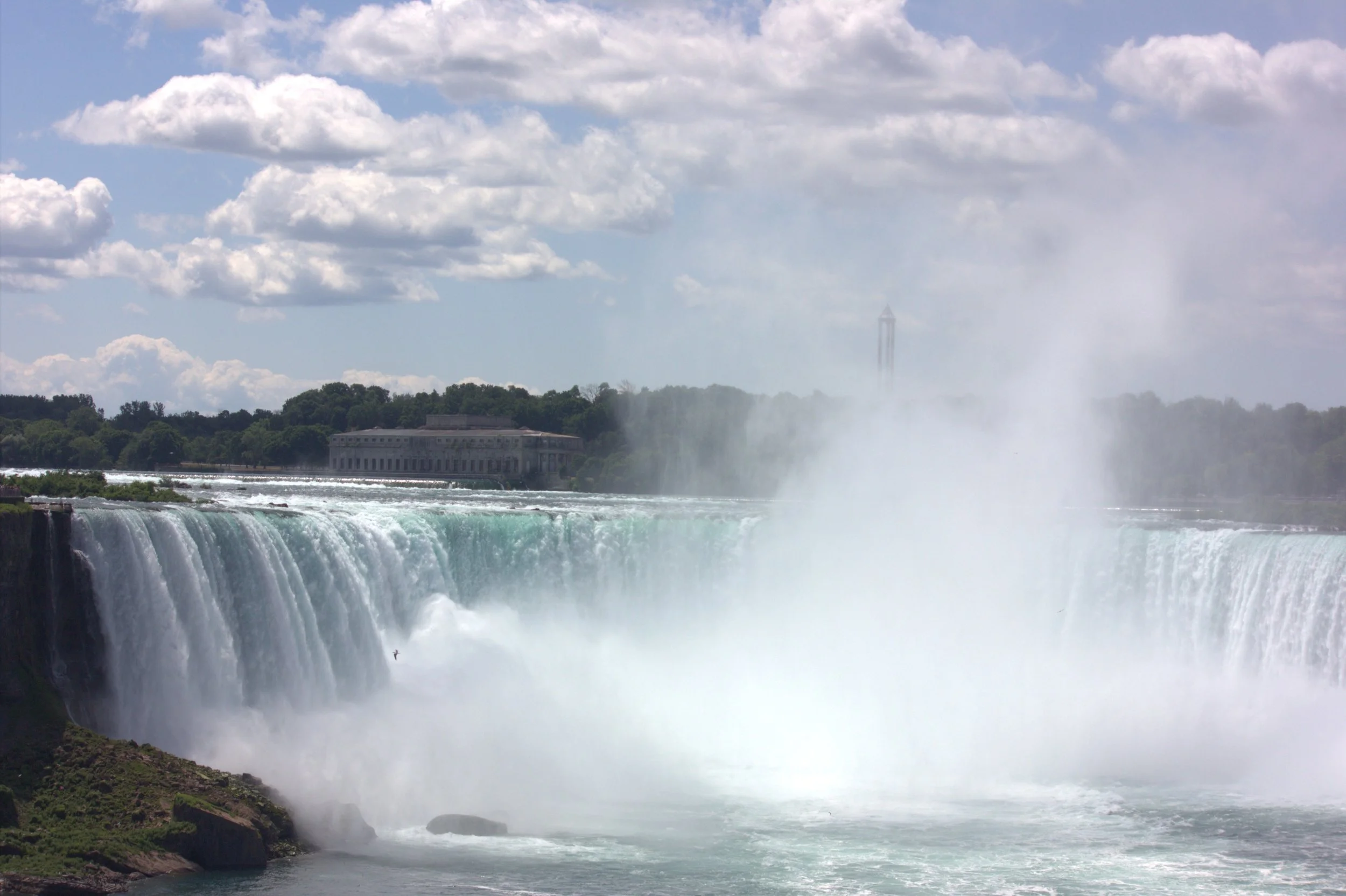 View of Niagara Falls with water cascading over the edge, mist rising, a building on the far side, and a partly cloudy sky.