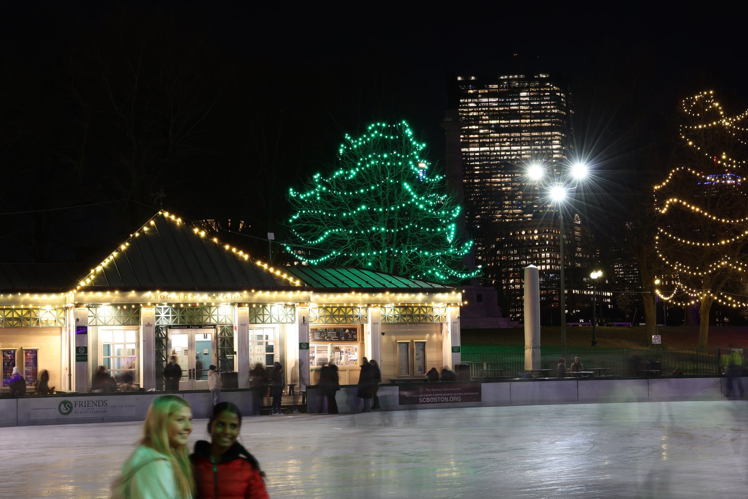 Ice skaters at an outdoor rink with festive holiday lights and a building with illuminated windows in the background at night.
