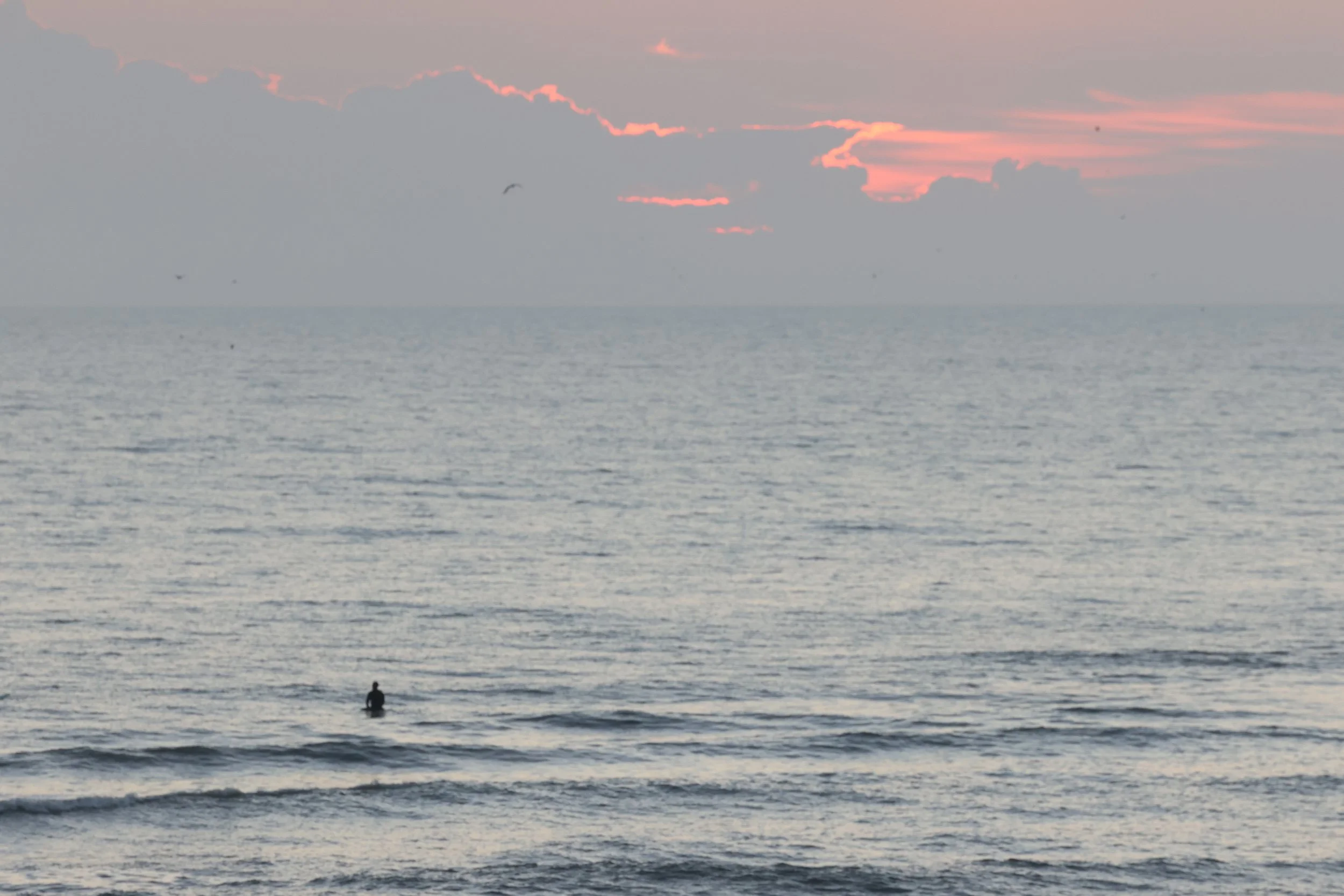 A person stands in the ocean waves during sunset, with the sky tinged with soft pink and orange hues and clouds partially obscuring the horizon.